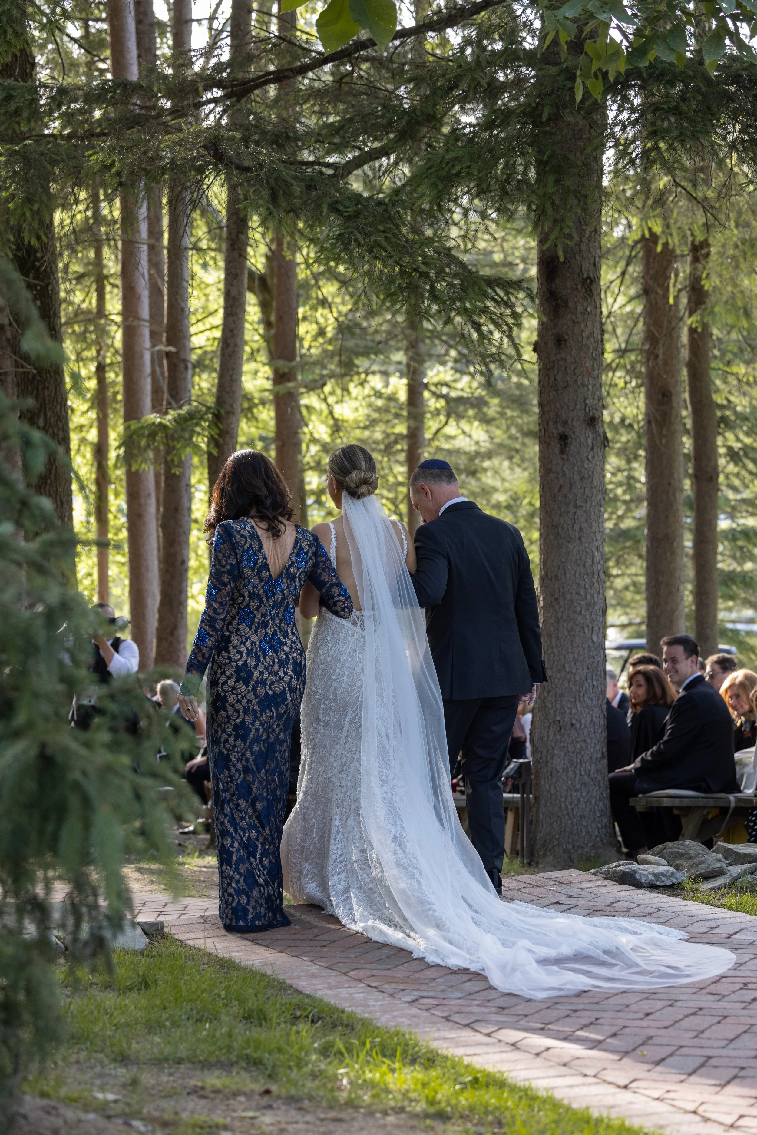 A bride walking down the aisle on her wedding day, accompanied by two women, in an outdoor forest setting with seated guests in the background.