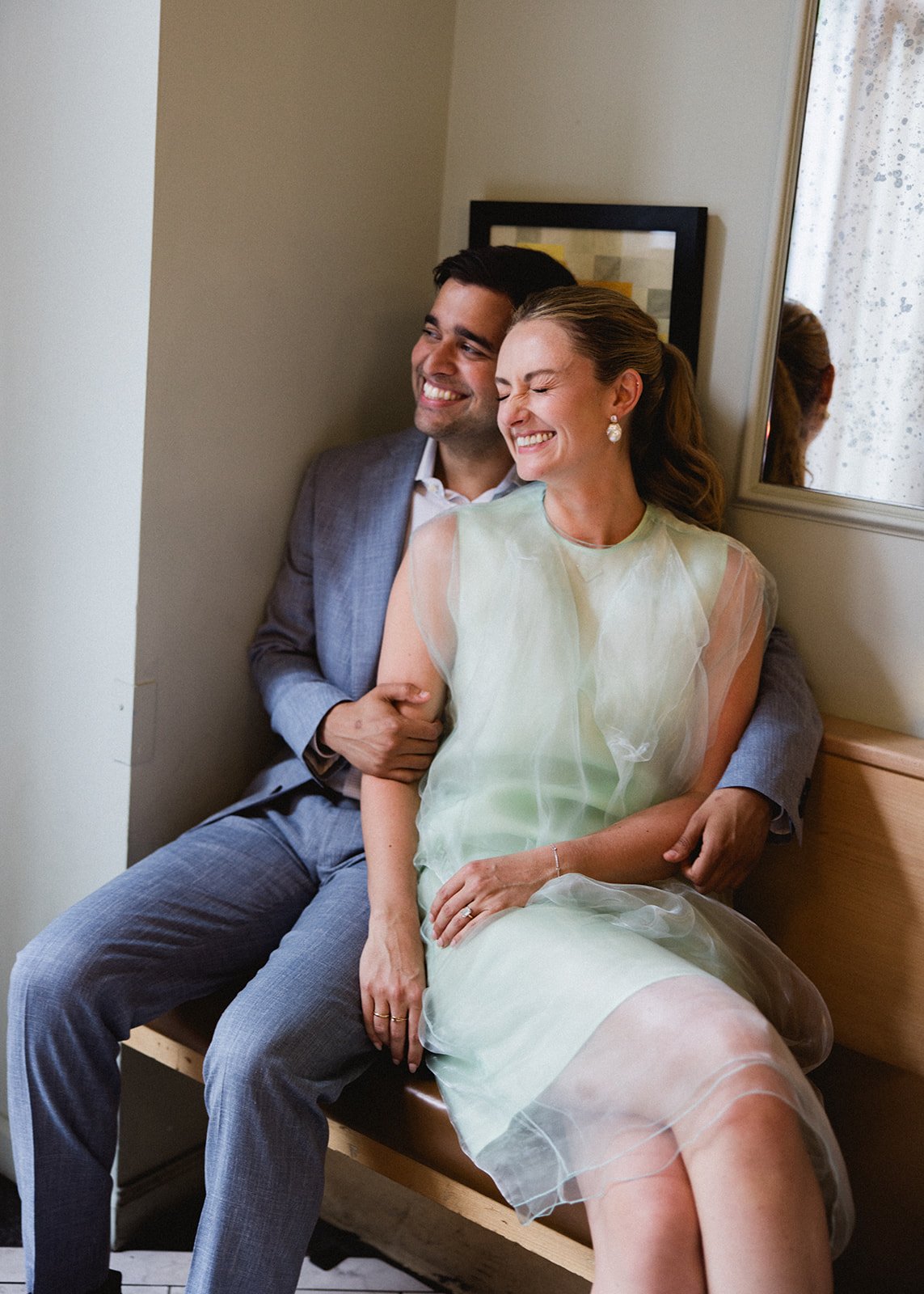 A happy couple sitting on a wooden bench, laughing and embracing each other in a cozy room with a framed picture and window with curtains