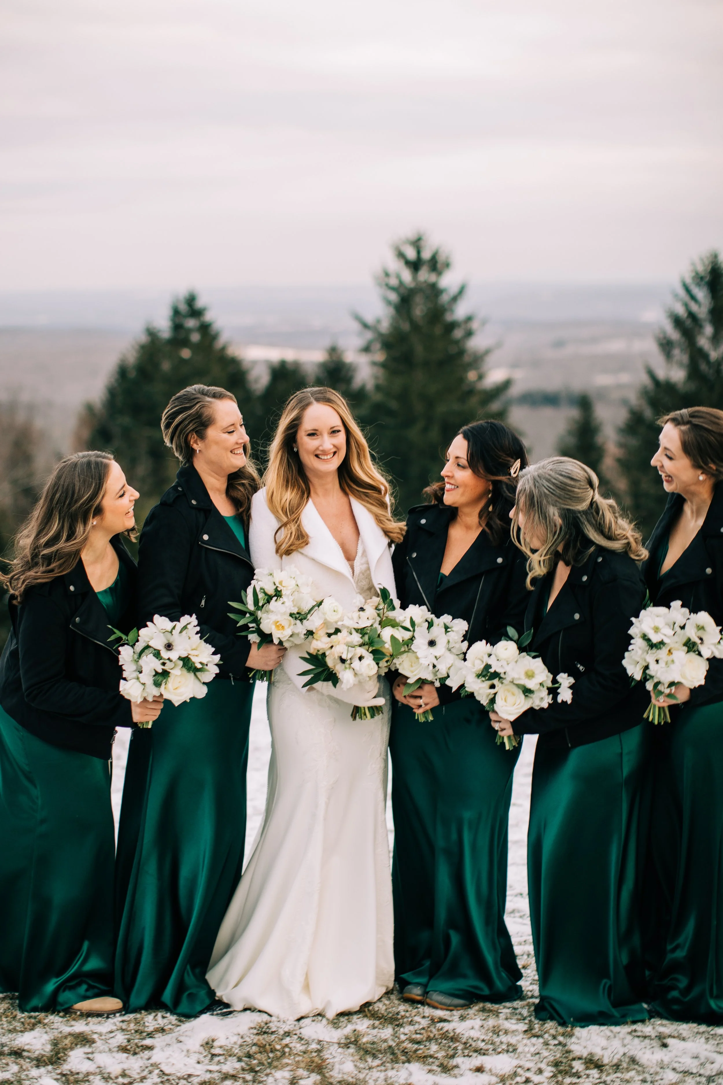 Bride in white dress with bridesmaids in dark green dresses and black jackets holding white bouquets outdoors.
