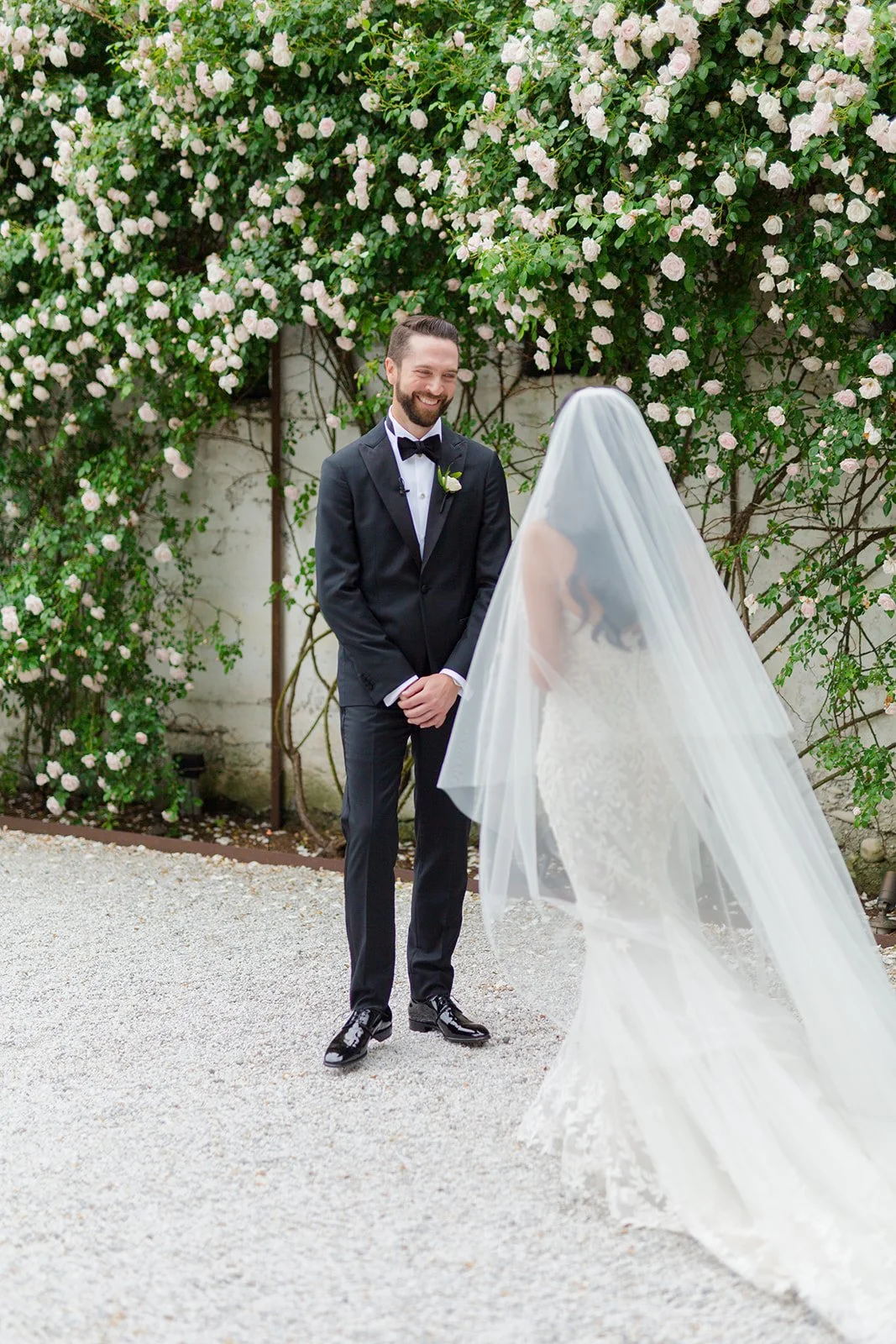 A groom wearing a black tuxedo with a bow tie, smiling at a bride with a veil, standing outdoors in front of pink and white roses