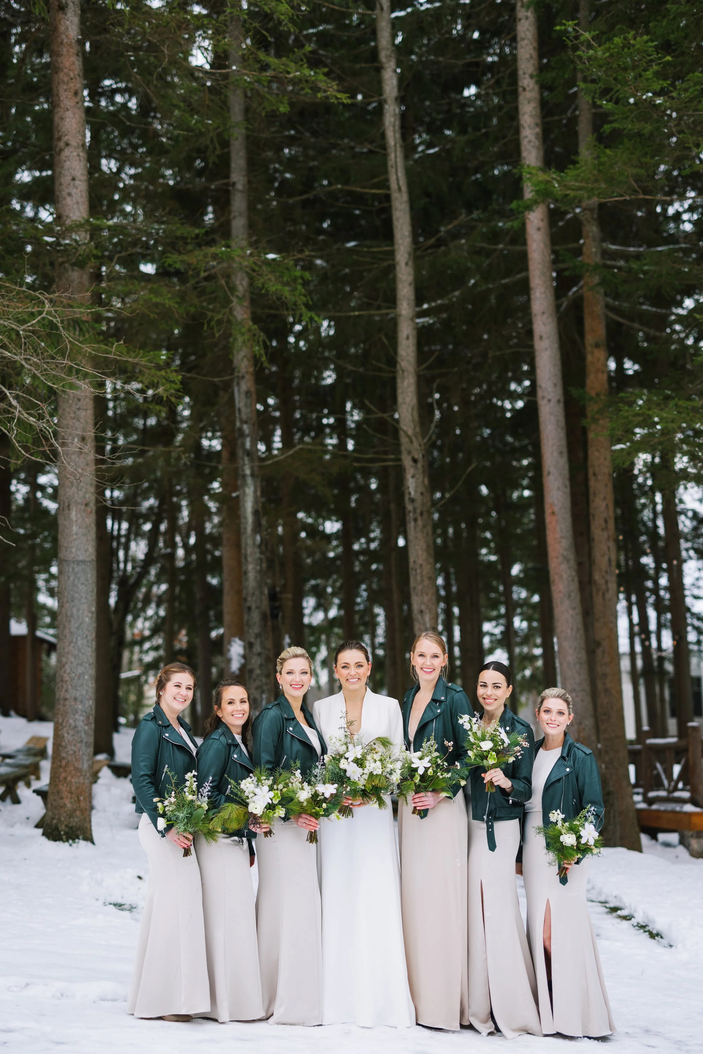 Bride and bridesmaids standing outdoors on snow, holding flower bouquets, with tall pine trees in the background.