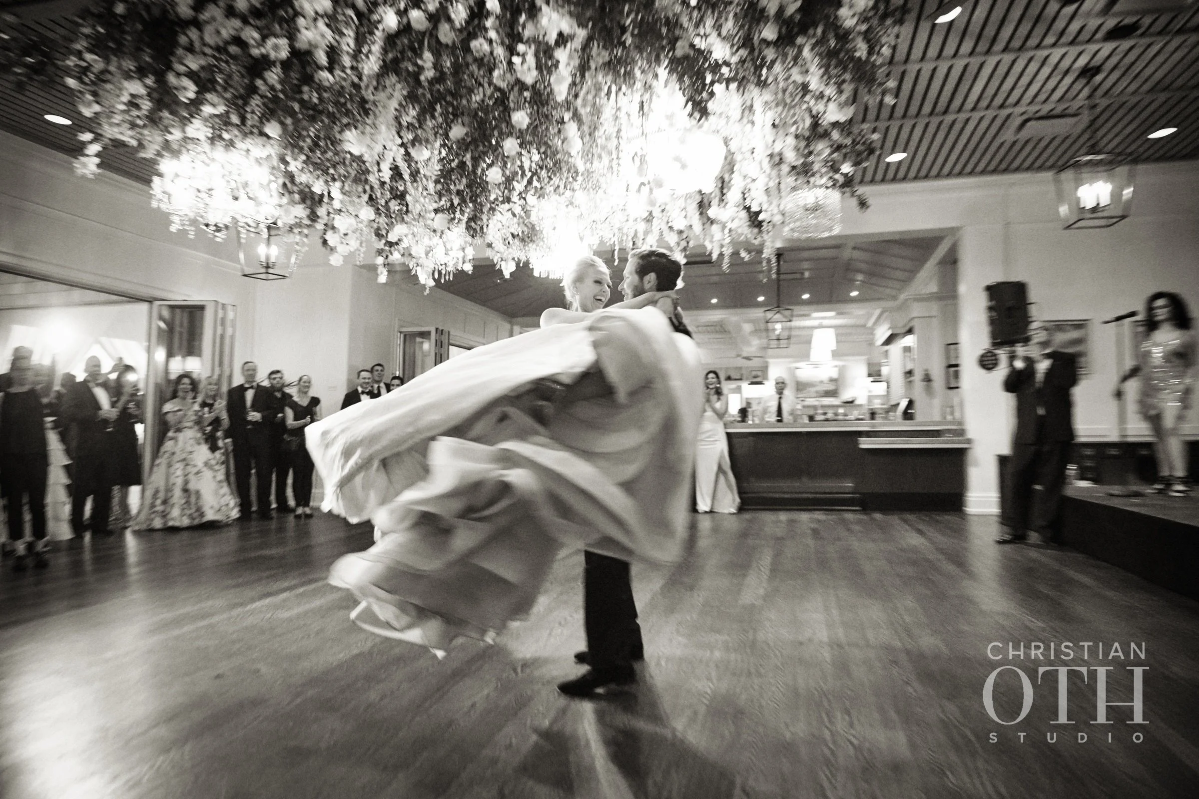 A bride and groom dance at their wedding reception, with guests watching in the background and a floral decoration hanging from the ceiling.