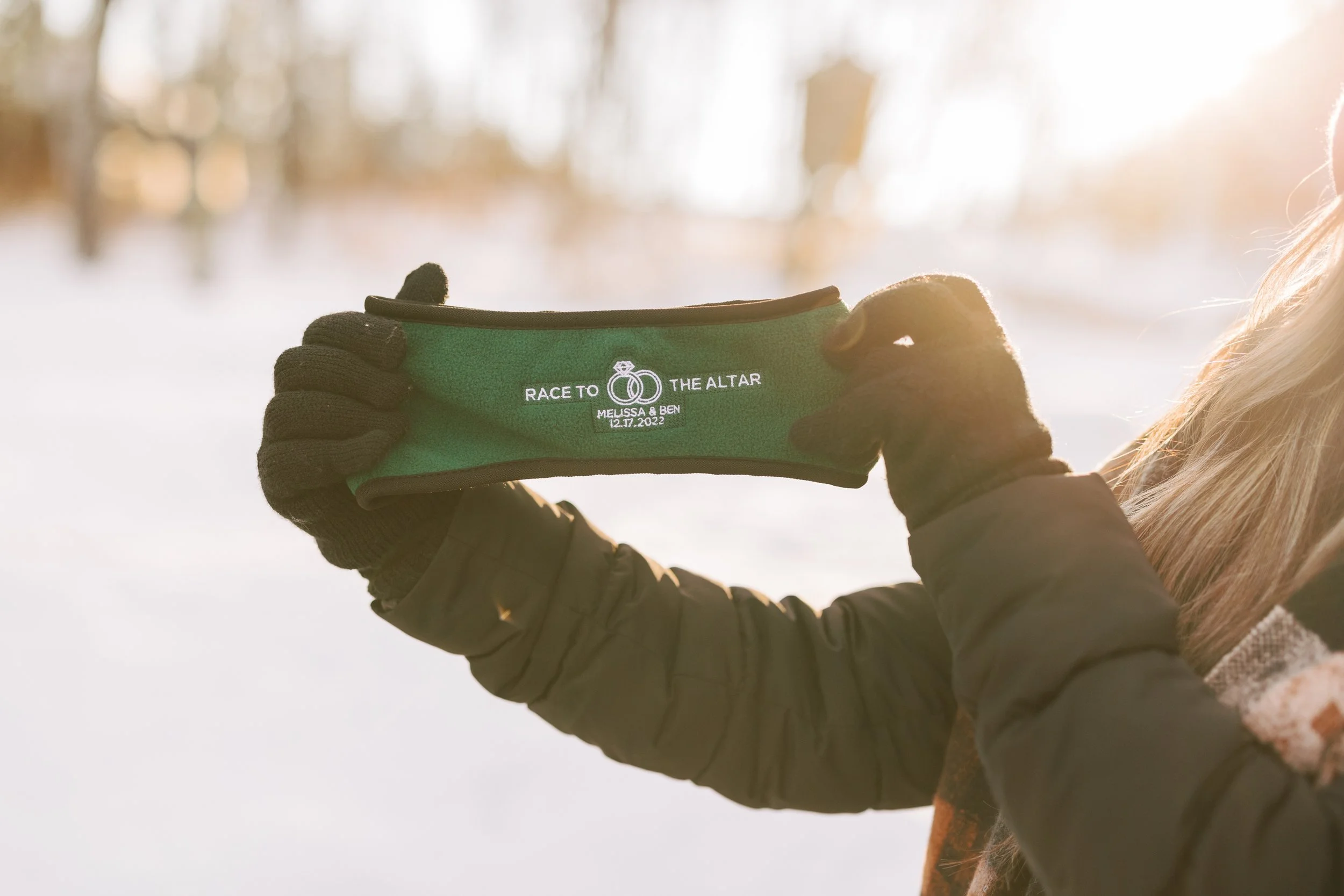 Person holding a green fabric band with white text that reads 'Race to the Altar, Melissa & Ben, 12.17.2022' in a snowy outdoor setting, wearing black gloves and a winter coat.