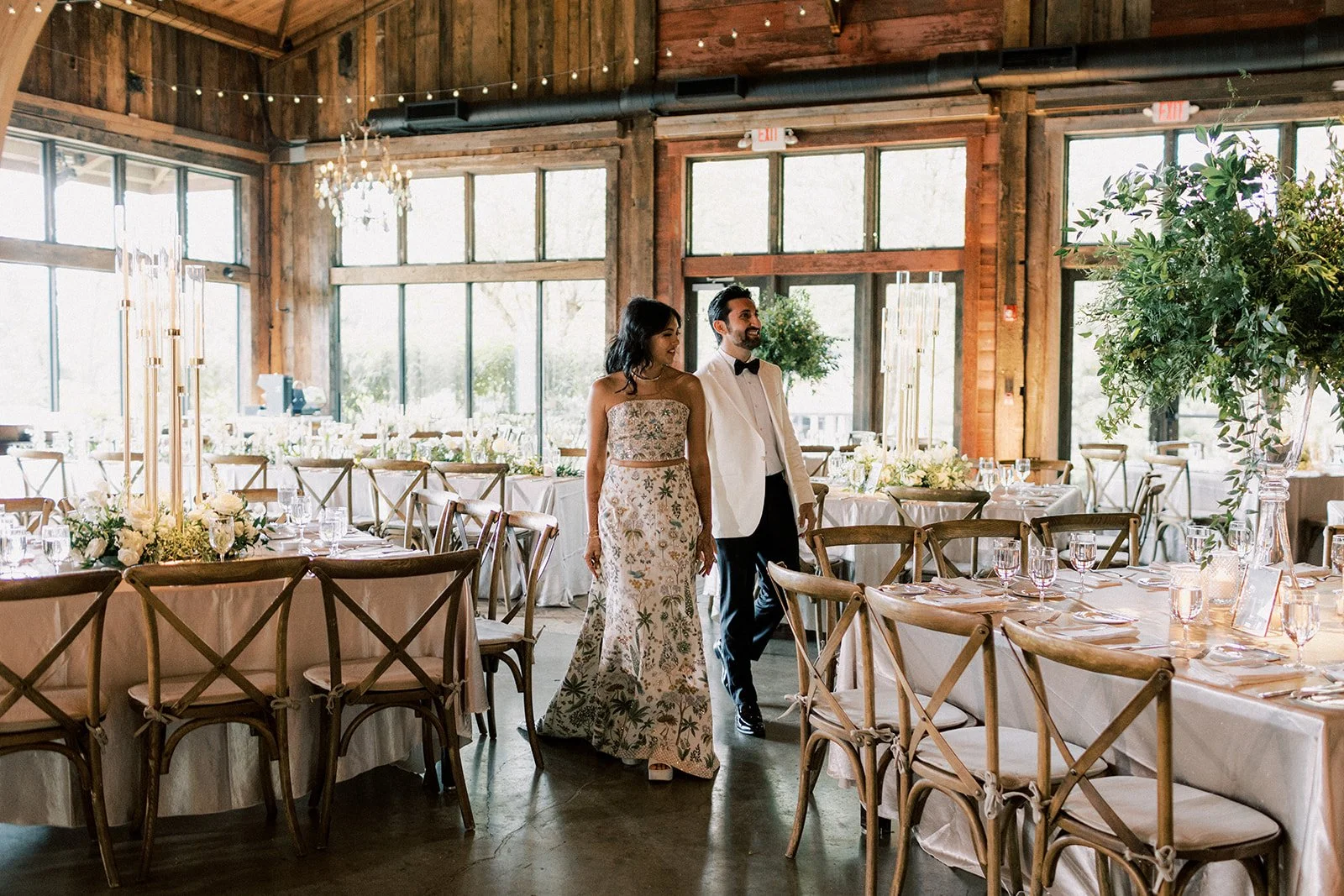 A couple dressed in formal attire walking inside a rustic wedding venue with wooden walls, large windows, and decorated tables.