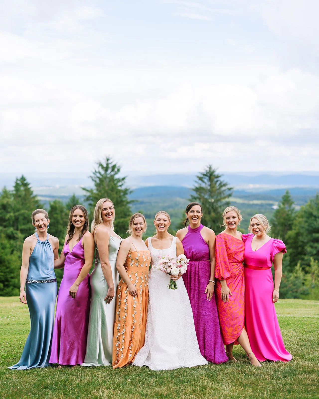 Group of women standing outdoors, dressed in colorful dresses, with one woman in a white wedding gown holding a bouquet, smiling against a backdrop of green trees and a cloudy sky.
