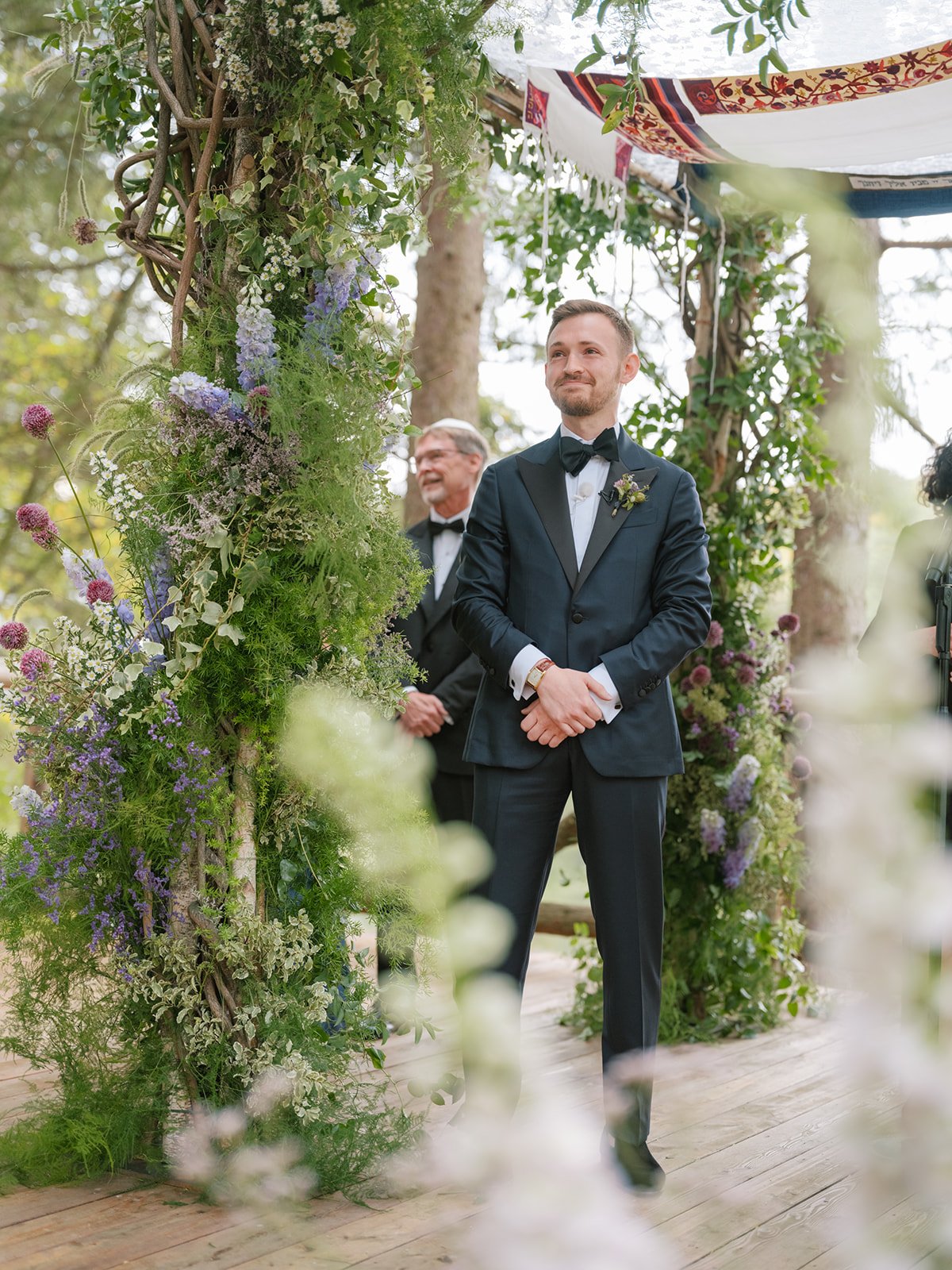 Groom in a dark blue tuxedo with a black bow tie standing at a wedding altar decorated with green plants and flowers outdoors, smiling and looking forward, with a man in a tuxedo smiling in the background.