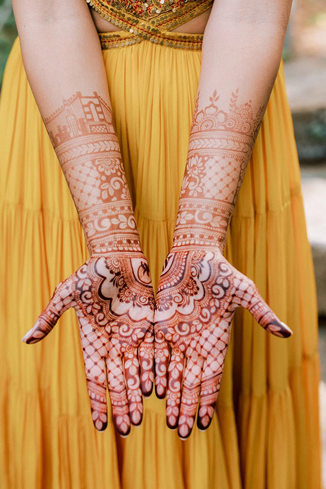 A person wearing a flowing yellow dress with ornate embroidery and jewelry, holding out their hands decorated with intricate henna designs.