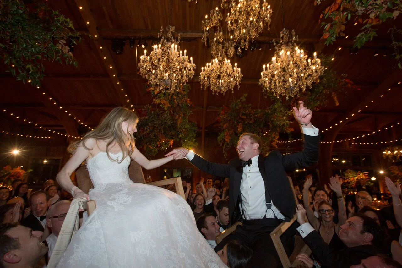 A wedding celebration with a bride in a white gown and a groom in a tuxedo, sitting on chairs, holding hands, and smiling. The scene is illuminated by chandeliers and string lights, with guests surrounding them, enjoying the event.