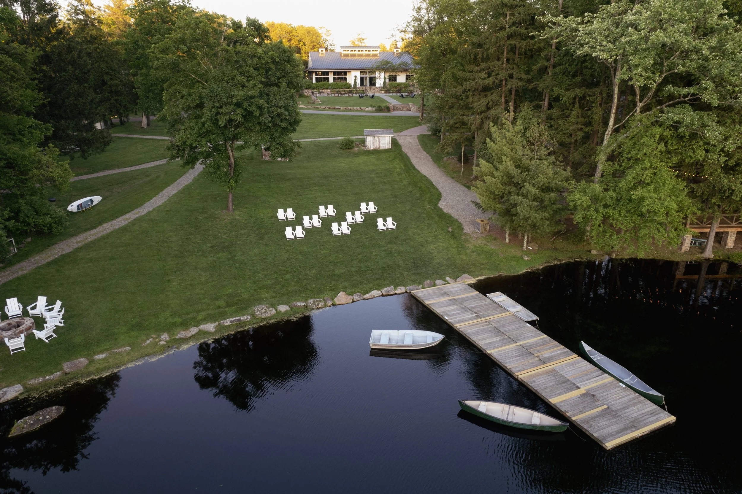 Aerial view of a lakeside lawn with white Adirondack chairs, a dock with boats, surrounded by trees, and a house in the background.
