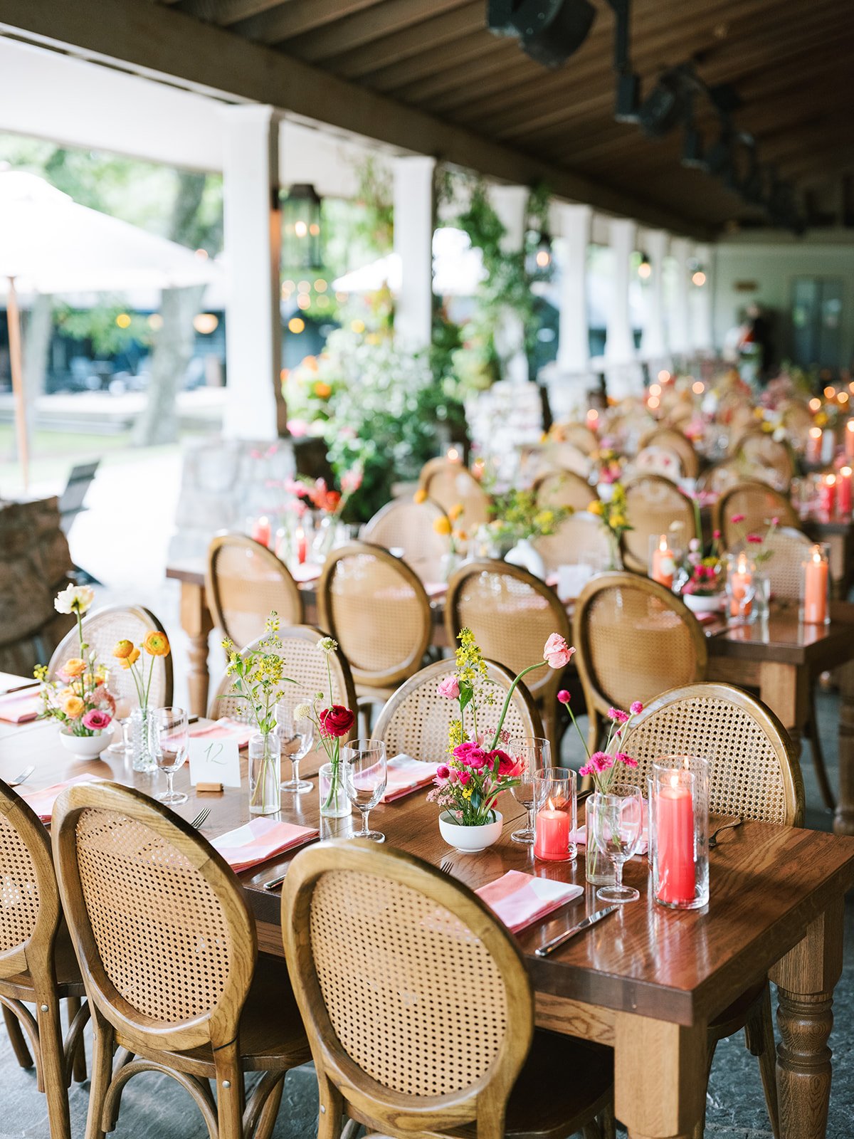 Decorated outdoor dining area with tables, chairs, pink and yellow floral centerpieces, lit candles in glass holders, and place settings for a celebration or event.