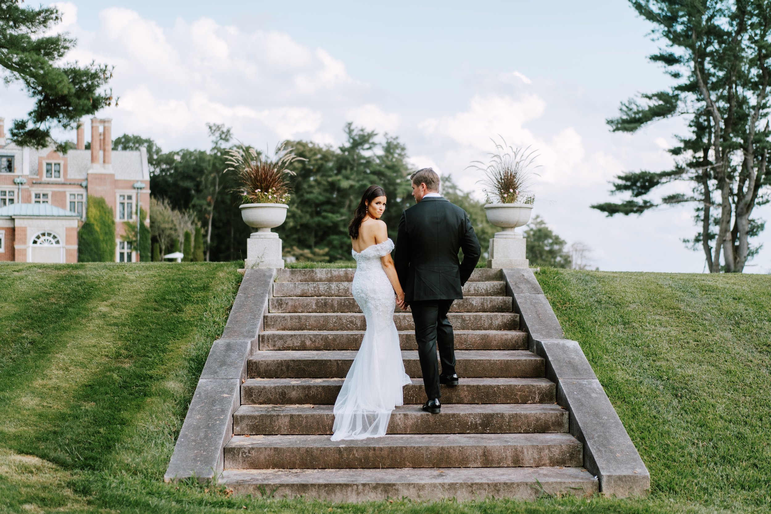 Bride and groom holding hands, walking up outdoor stone stairs, with large planters and a mansion in the background.