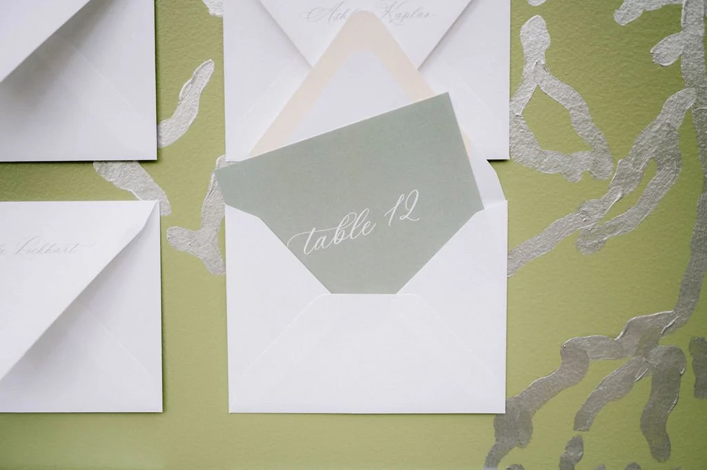 White and gray wedding place cards on a green and silver decorative background, with one card displaying "table 12".