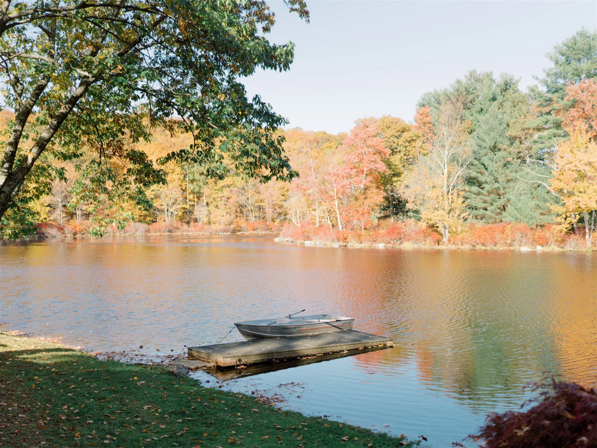 A peaceful lake scene with a small boat docked at a pier, surrounded by colorful autumn trees with leaves in shades of green, yellow, orange, and red.