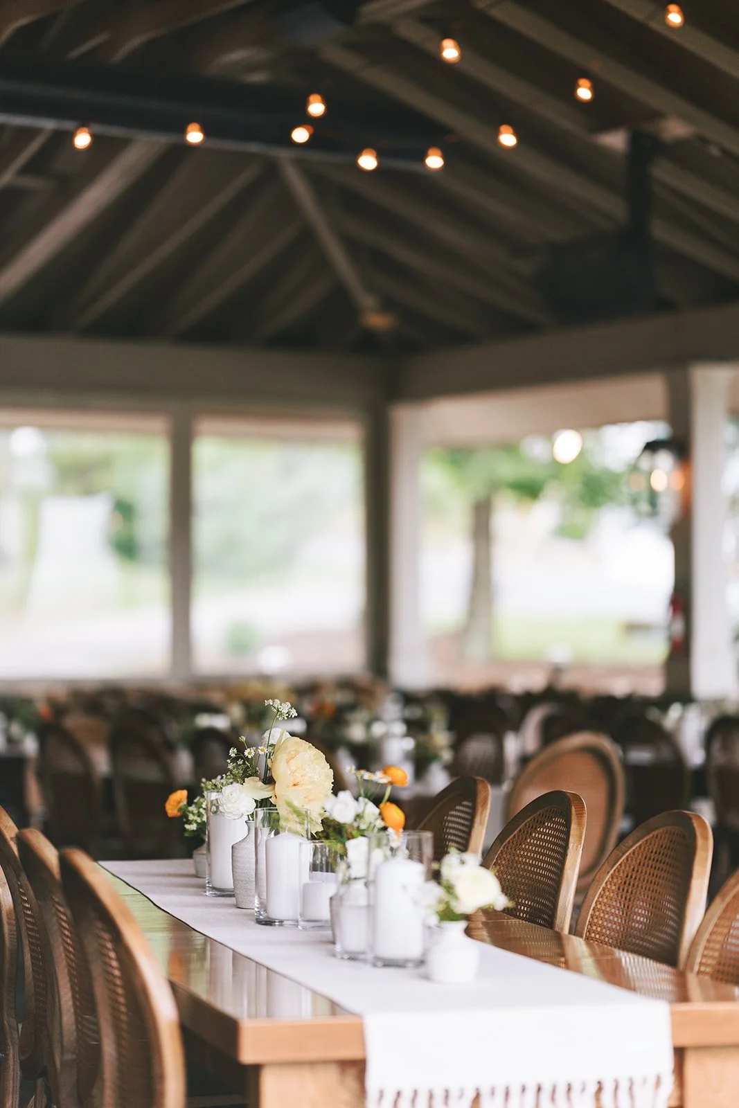 A long wooden banquet table decorated with white candles, white and cream flowers, and small orange accent flowers in white vases, set inside a rustic, open-air pavilion with string lights.