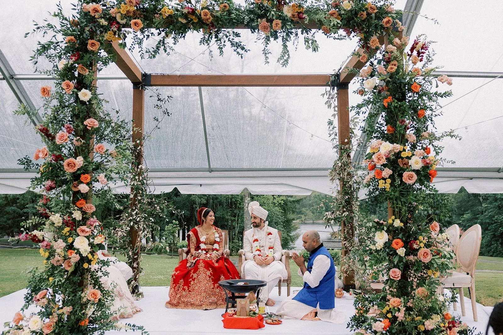 An outdoor wedding ceremony with a floral arch and a couple dressed in traditional Indian wedding attire, seated on chairs under the arch, with a priest performing a ritual in front of them.