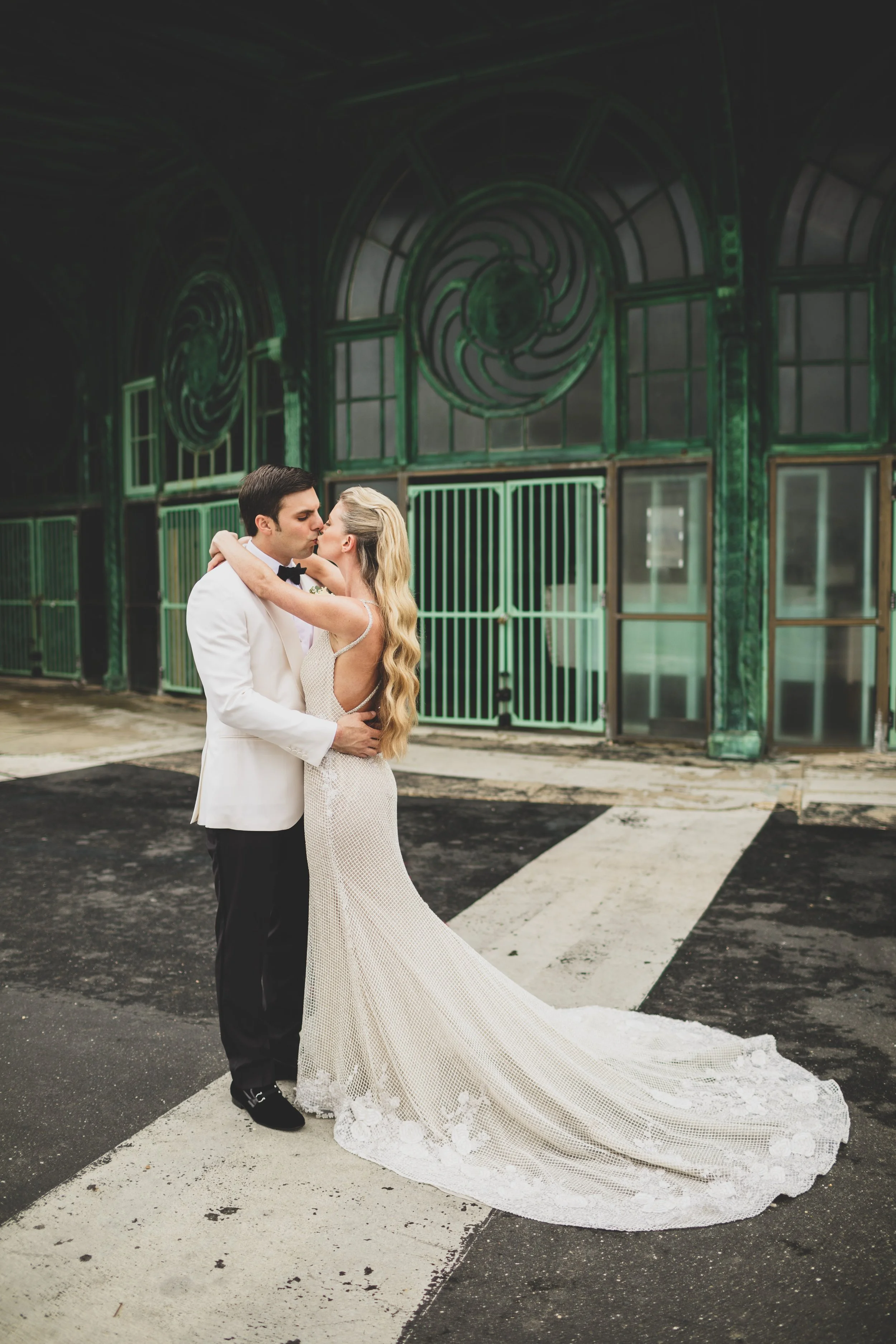 A bride and groom are kissing outdoors at night, with the bride in a long, white gown with a train, and the groom in a white tuxedo jacket and black pants, standing on a crosswalk with a green, art-deco building in the background.