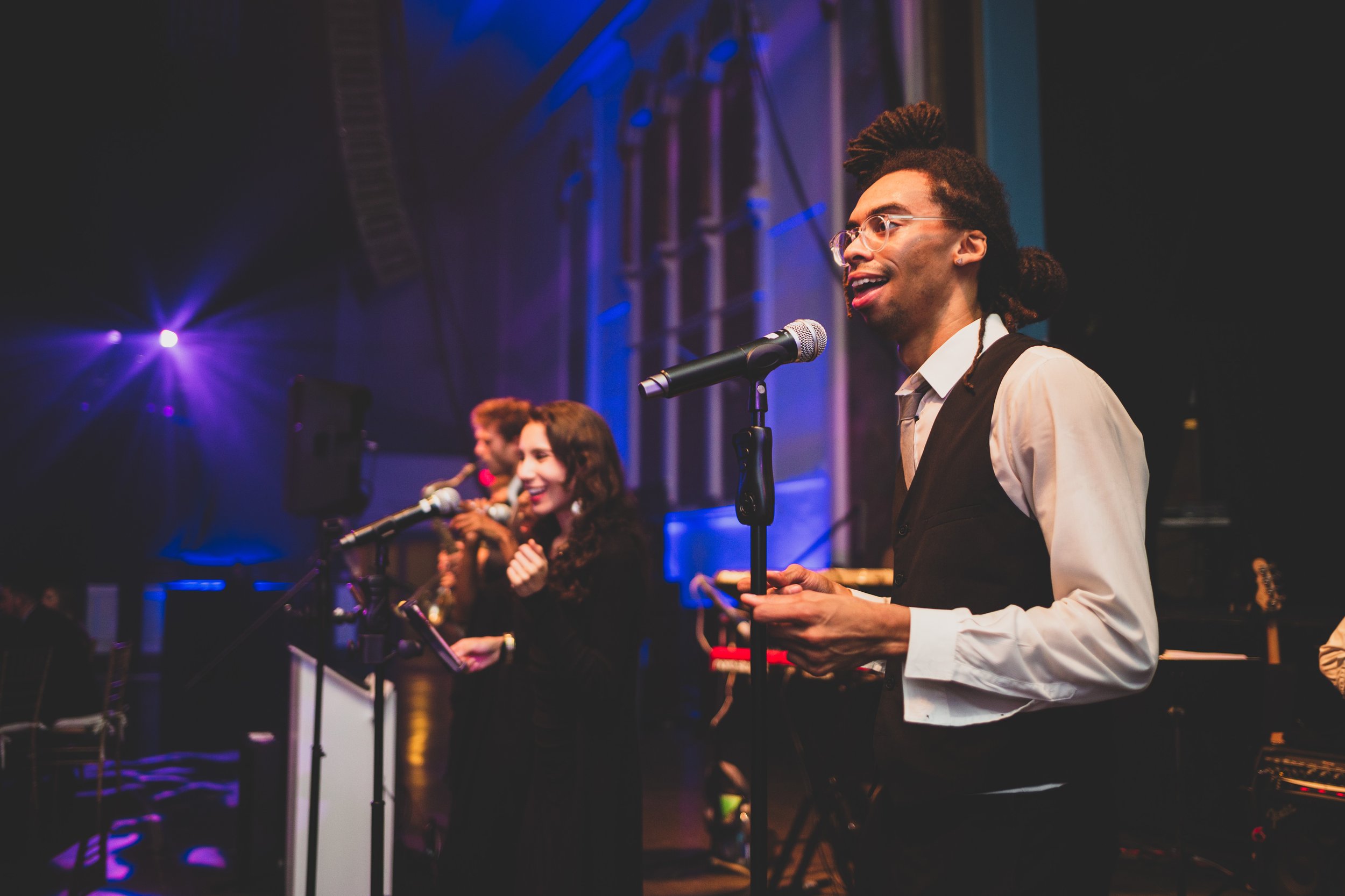 A group of performers singing or speaking on stage at a concert or event, with colorful stage lights and musical equipment visible.