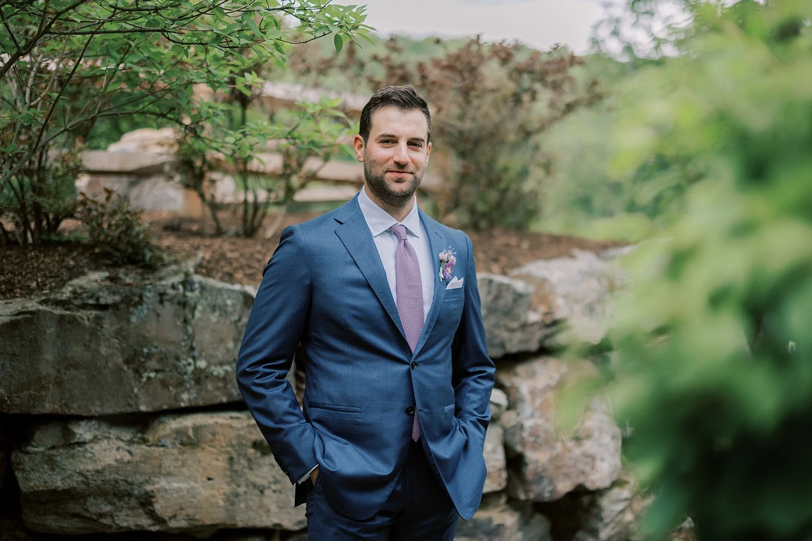 A man in a blue suit with a pink tie and boutonniere standing outdoors in front of rocks and greenery.