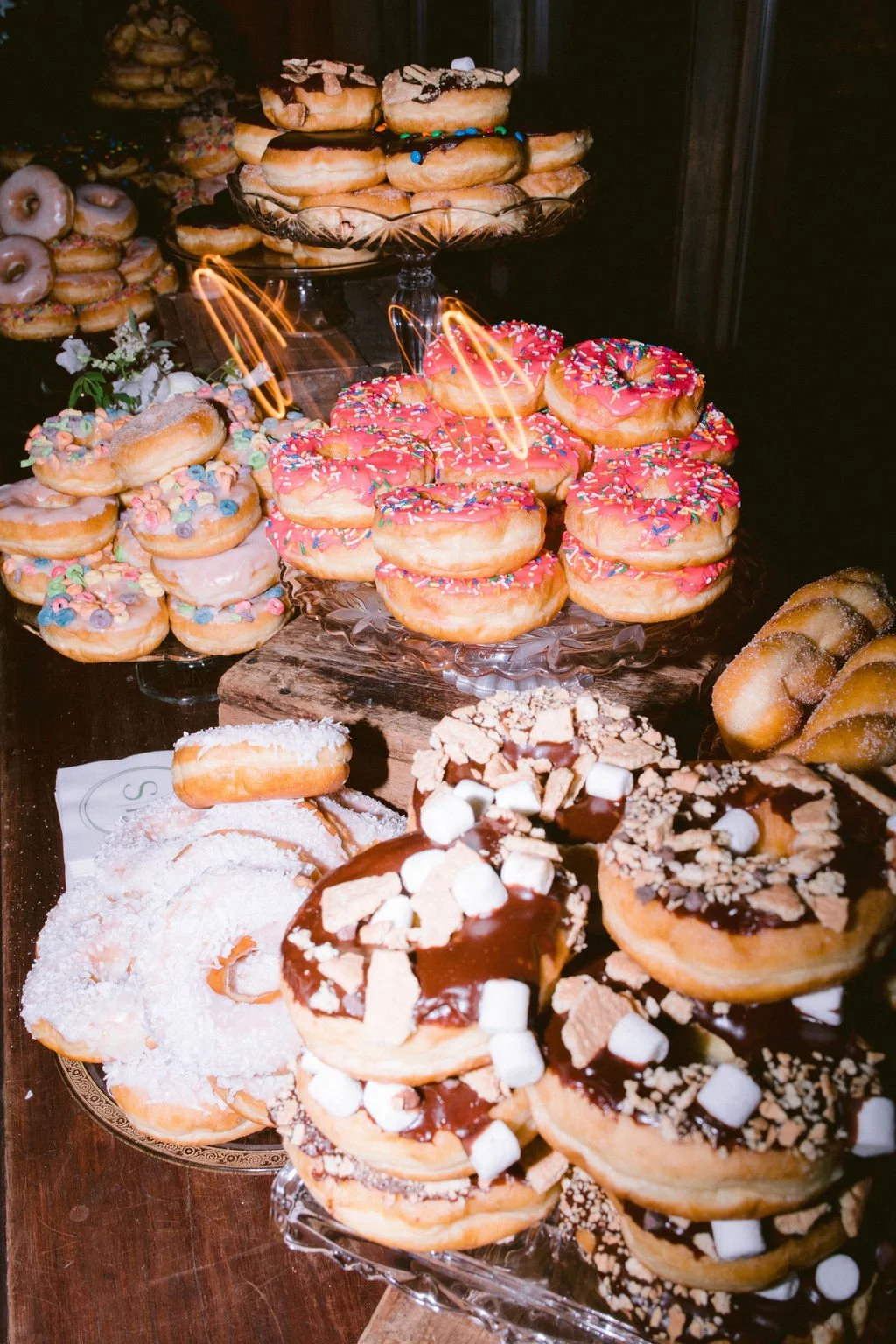 Assorted donuts with colorful sprinkles, chocolate, marshmallows, coconut flakes, and crushed cookies on display, some topped with icing and candies, arranged on glass and wooden stands.