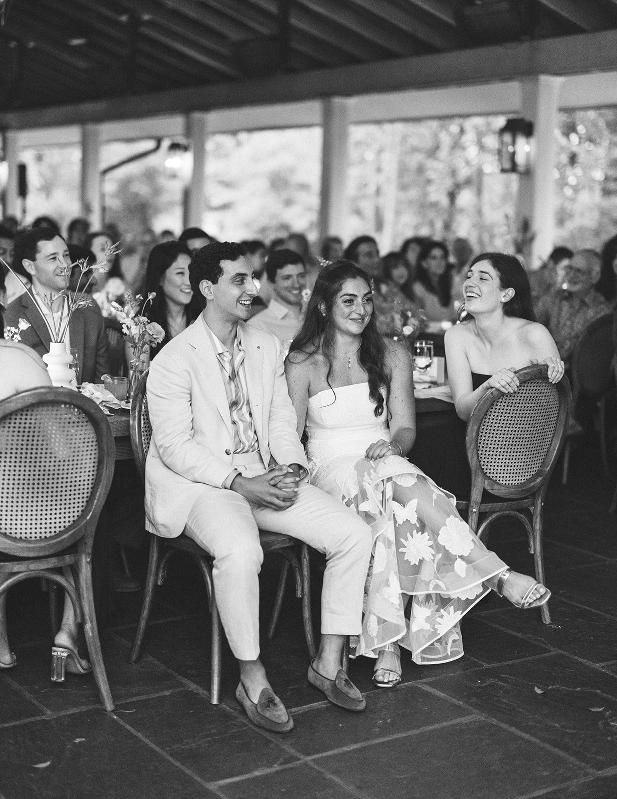 People sitting at a table during a celebration, with some smiling and laughing, in a decorated indoor setting.