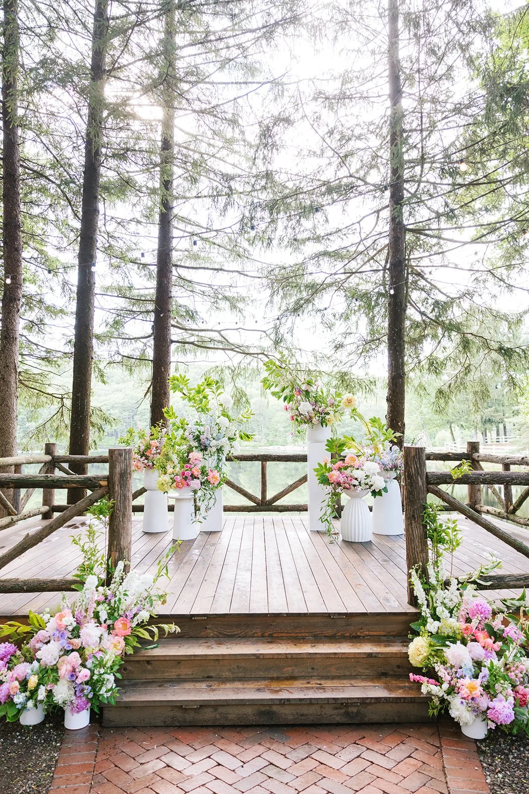Outdoor wedding ceremony altar on wooden platform, decorated with pink, white, and purple flowers in white vases, surrounded by a rustic wooden railing and tall trees in the background.
