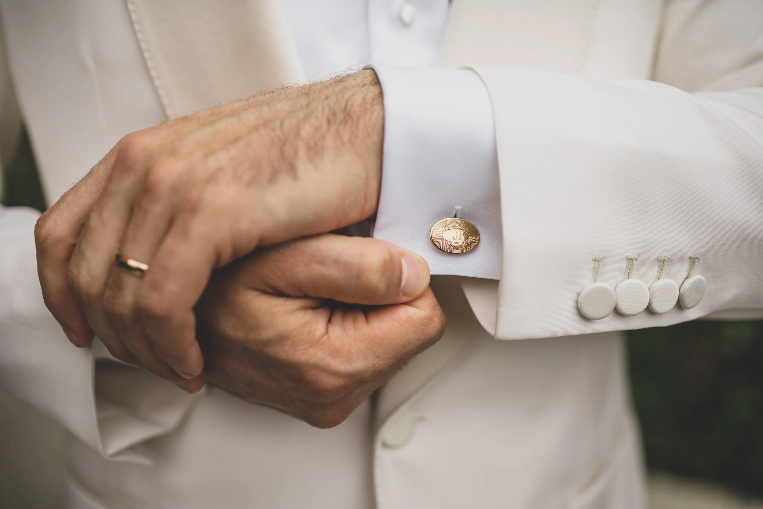 Close-up of a man adjusting his white tuxedo cuff with gold cufflink, wearing a wedding band.