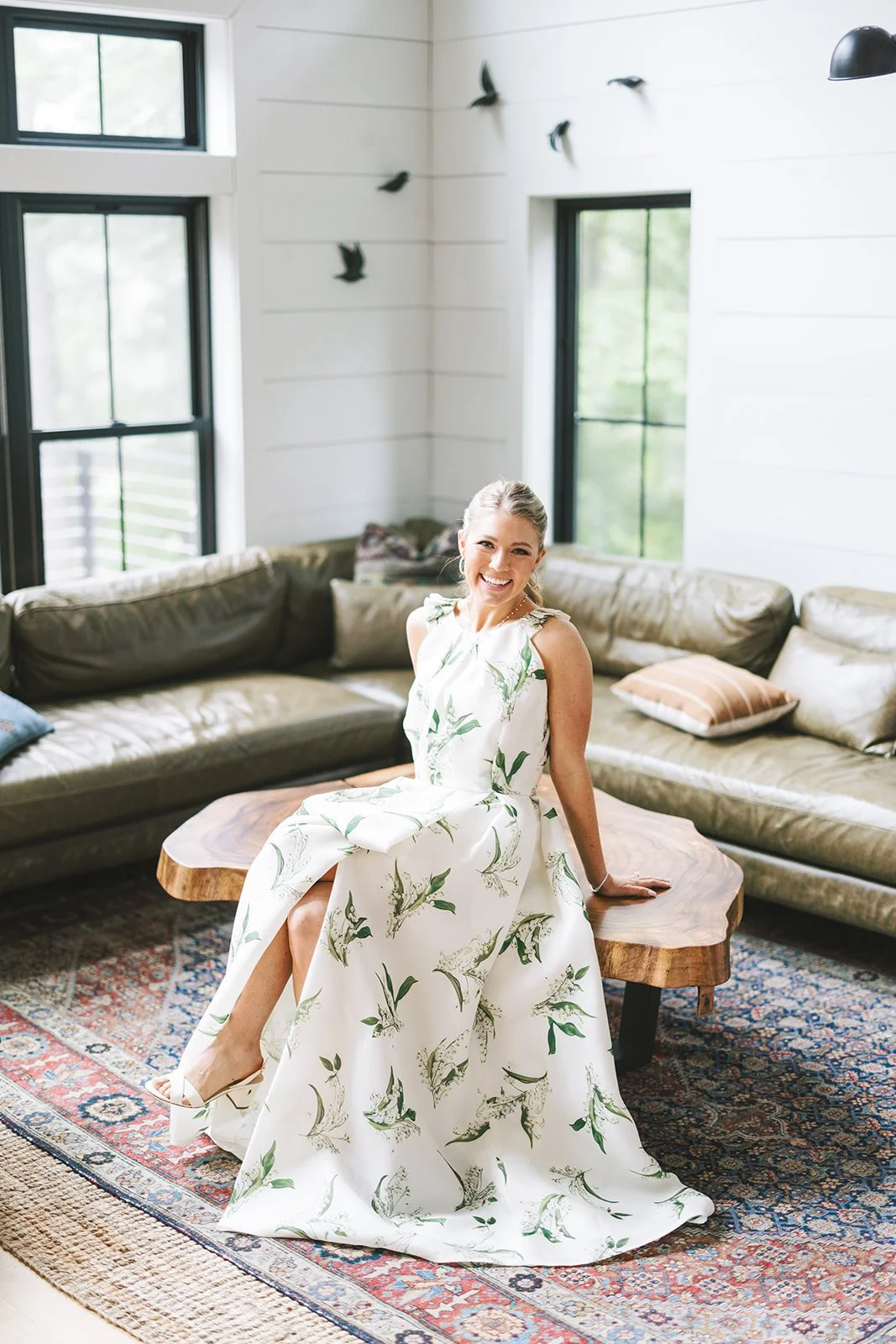 A woman in a white floral dress smiling while sitting on a wooden coffee table in a bright living room with large windows, beige sectional sofa, and decorative pillows.