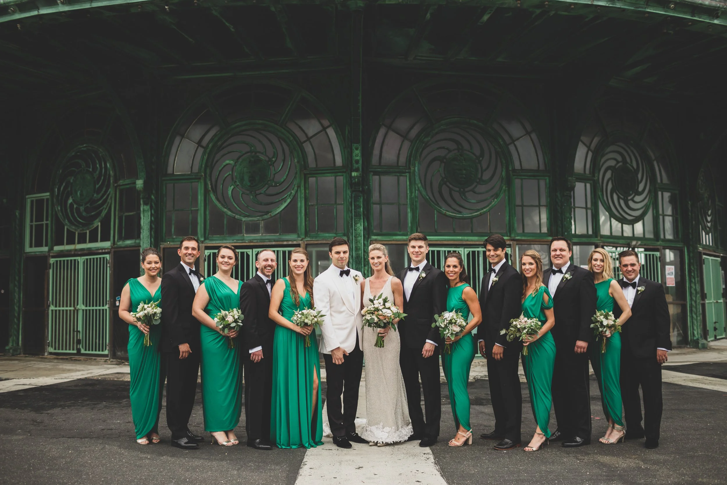 A wedding party of thirteen people standing in front of a large green industrial gate, with the bride and groom in the center. The bride wears a white wedding dress and holding a bouquet. The groom wears a white tuxedo jacket and black bow tie. Bride