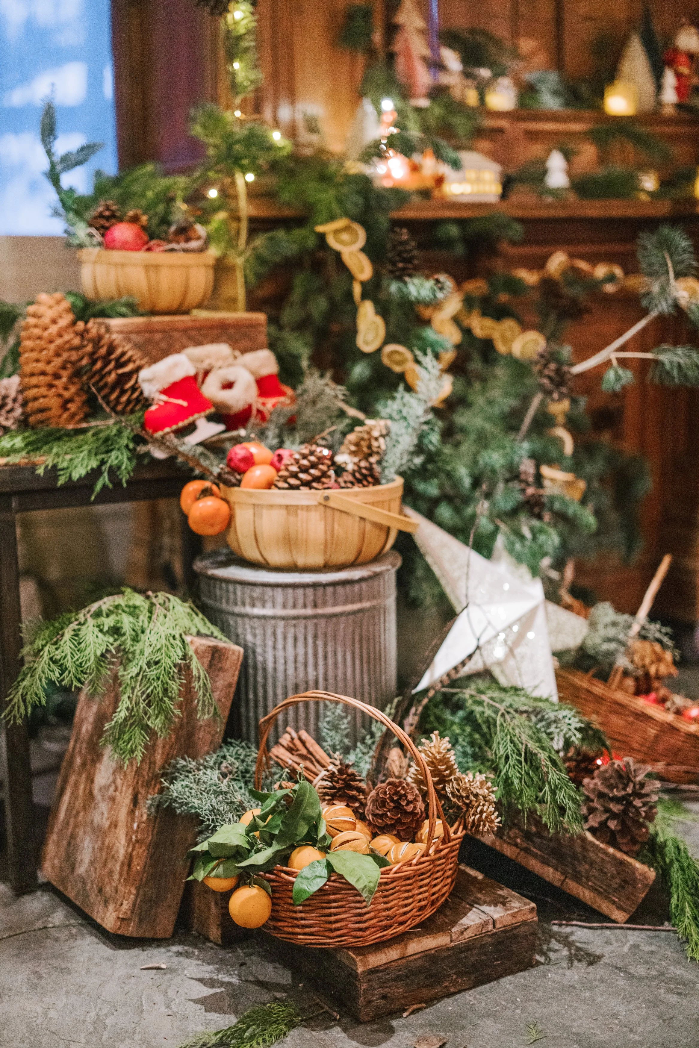 Decorative display of Christmas greenery, pinecones, and fruit in baskets and on wooden crates, with a lit star ornament and small festive figurines, arranged indoors with warm lighting.