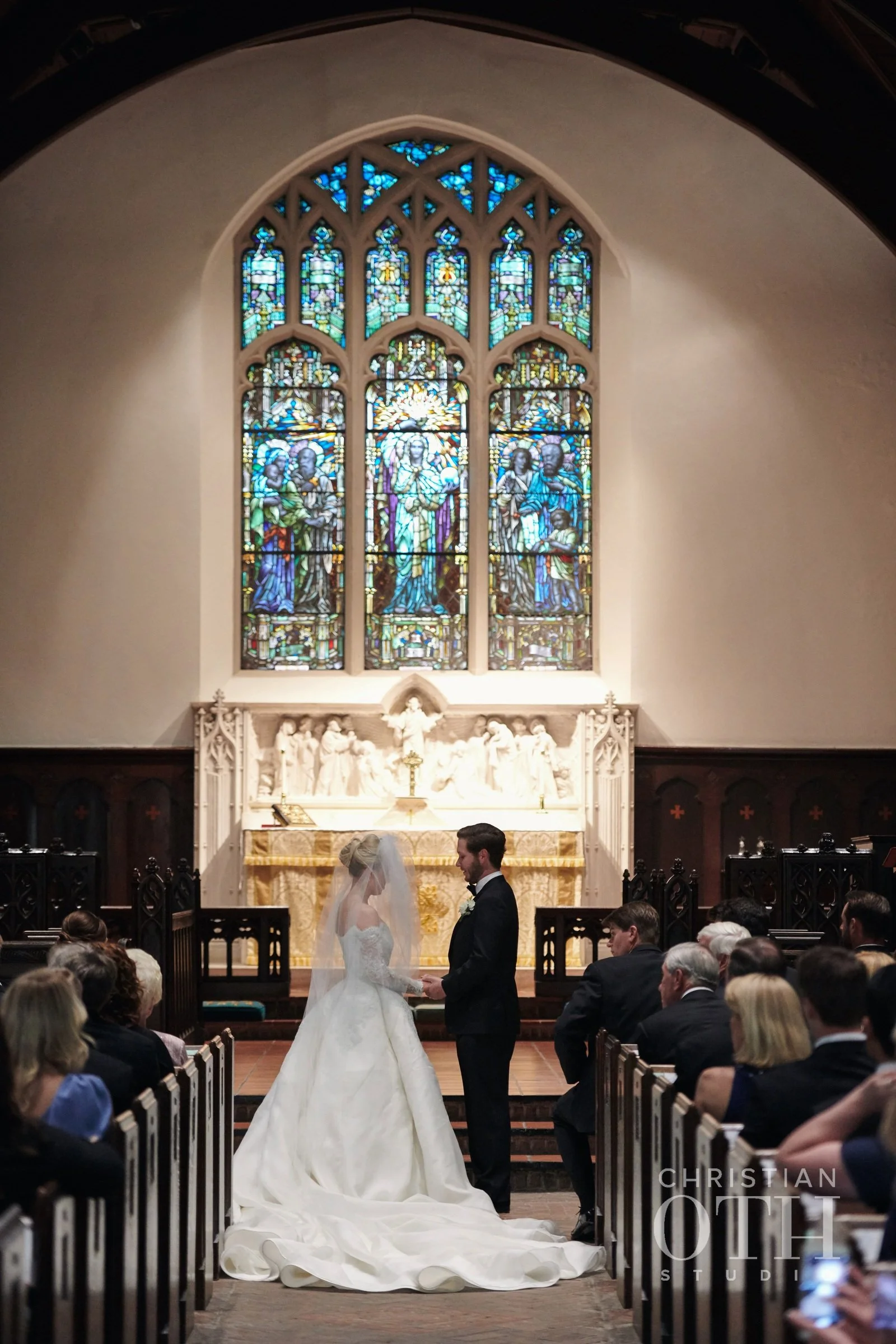 A bride and groom exchanging vows in a wedding ceremony inside a church with stained glass windows.