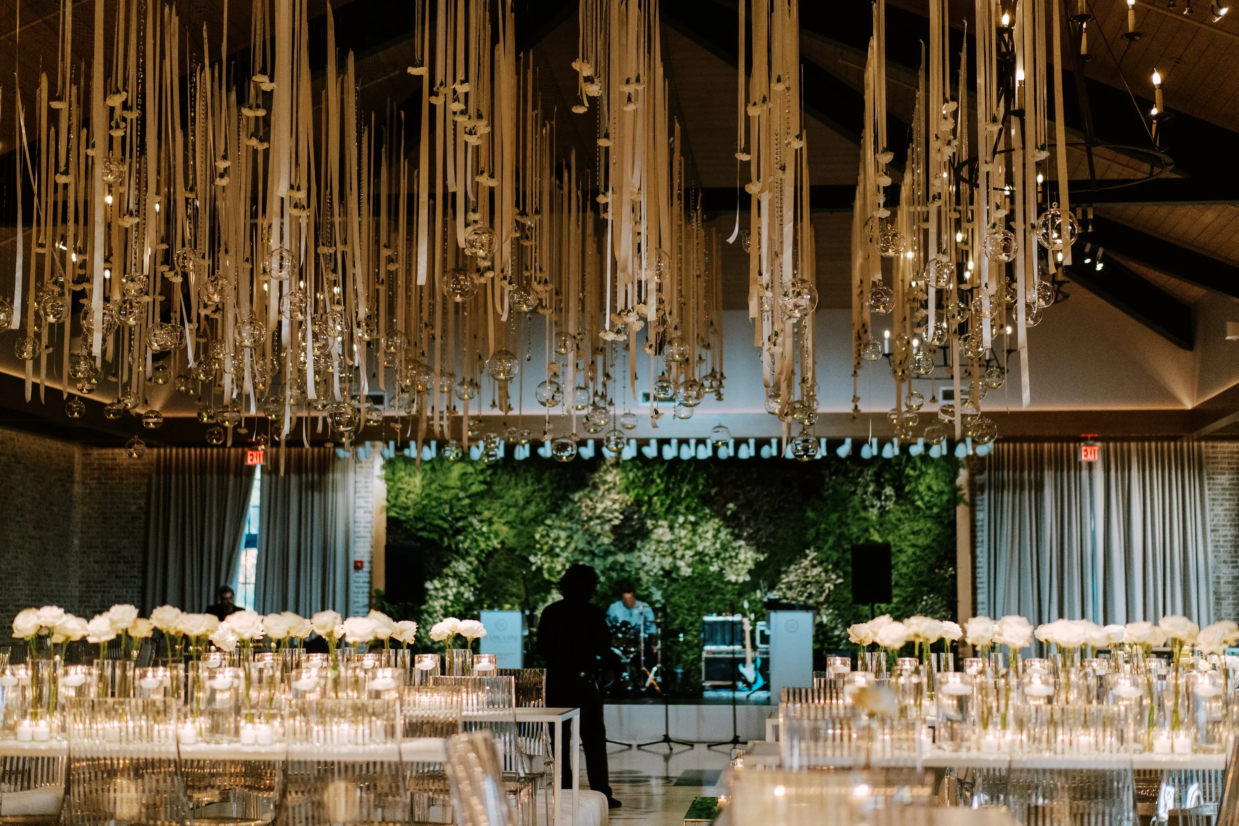 Elegant event space with long tables decorated with white flowers and candles, featuring a lush green backdrop, modern hanging glass globe chandeliers, and a small stage with musical instruments.