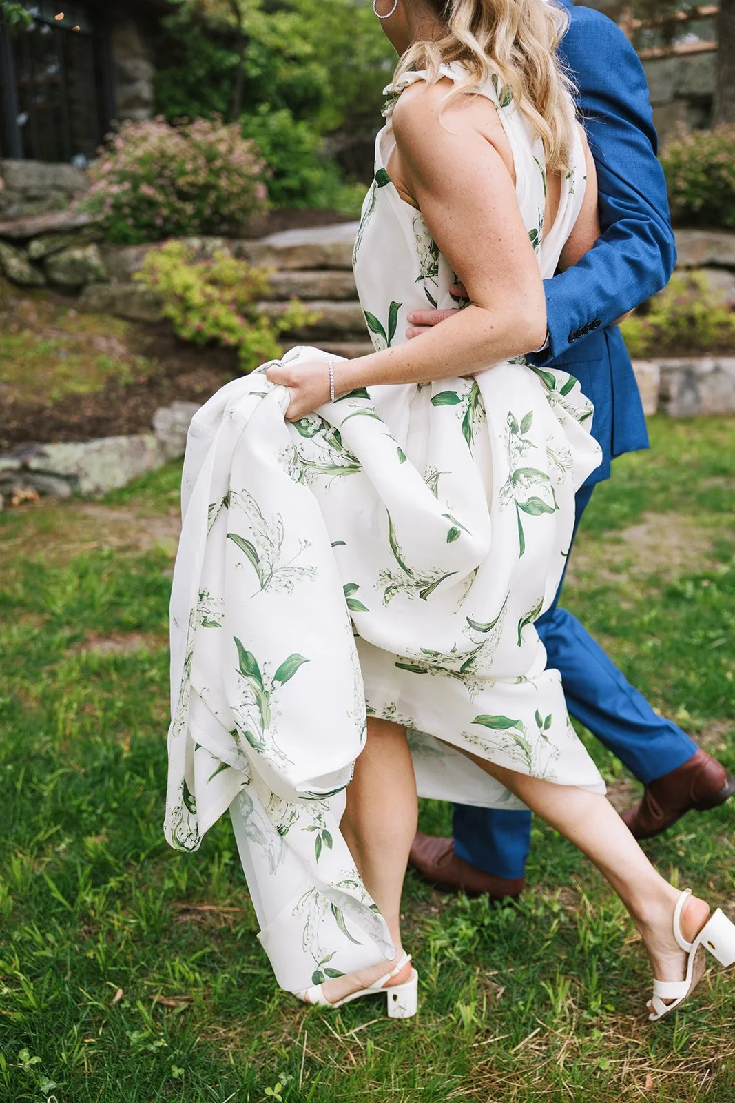 A woman in a white floral dress lifting her skirt as a man in a blue suit holds her from behind in a garden setting.