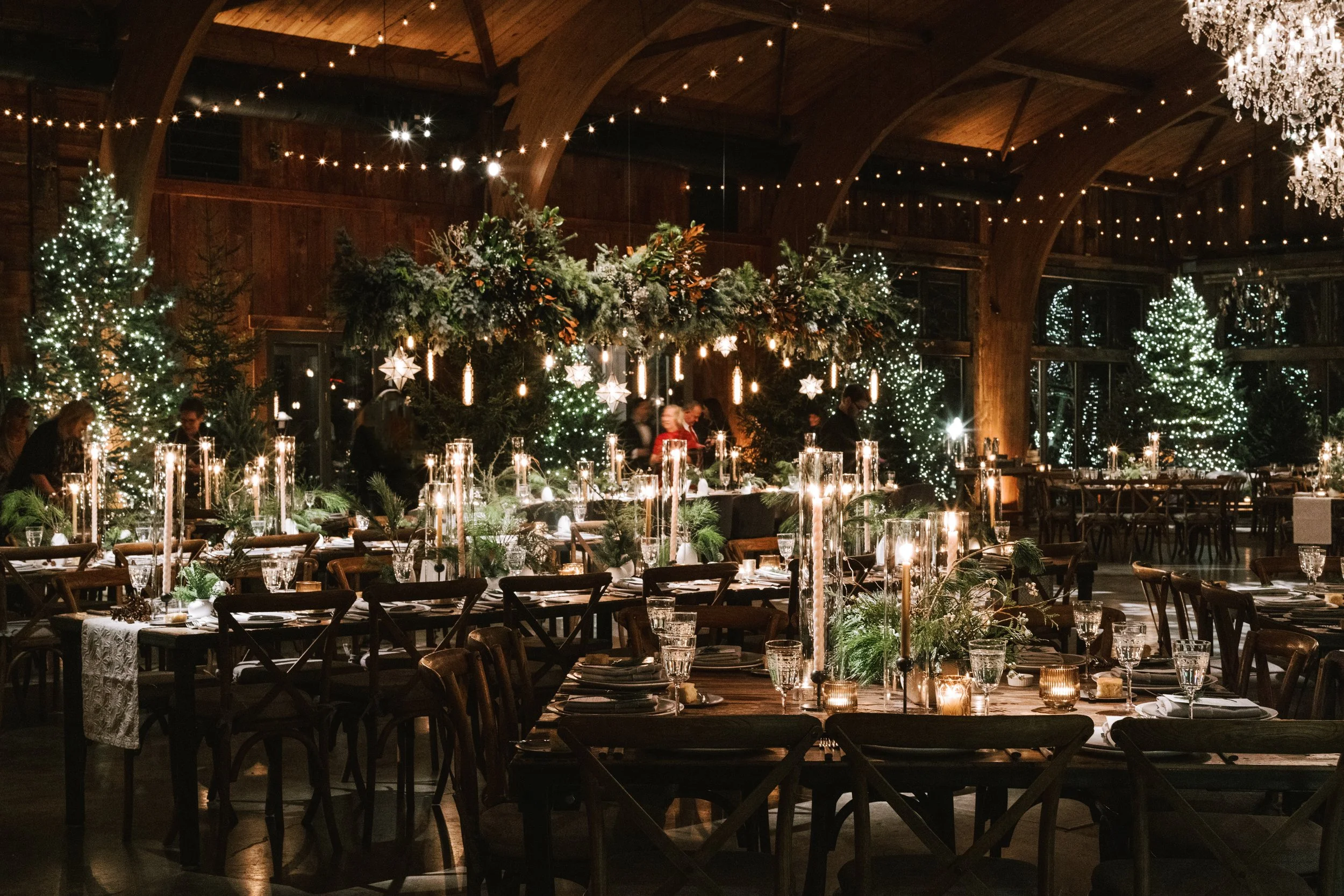 A festive indoor dining area decorated for Christmas, with tables set with candles, glassware, and greenery, Christmas trees with lights, and hanging star ornaments.