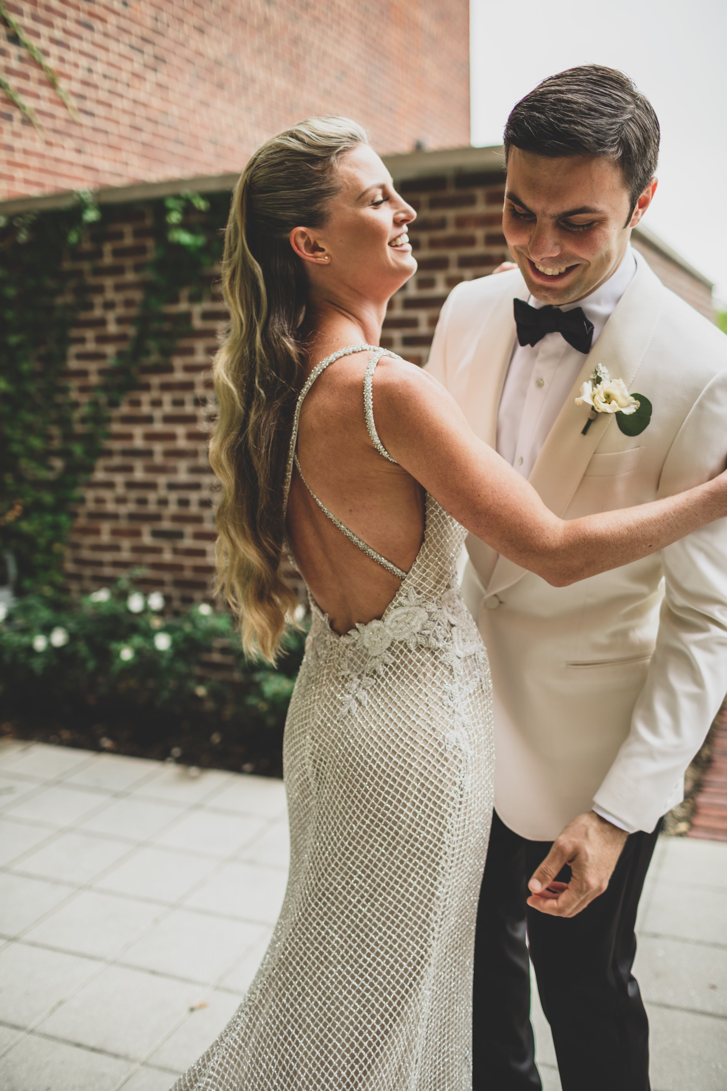 A bride and groom share a dance outdoors, smiling at each other, with a brick wall and greenery in the background.