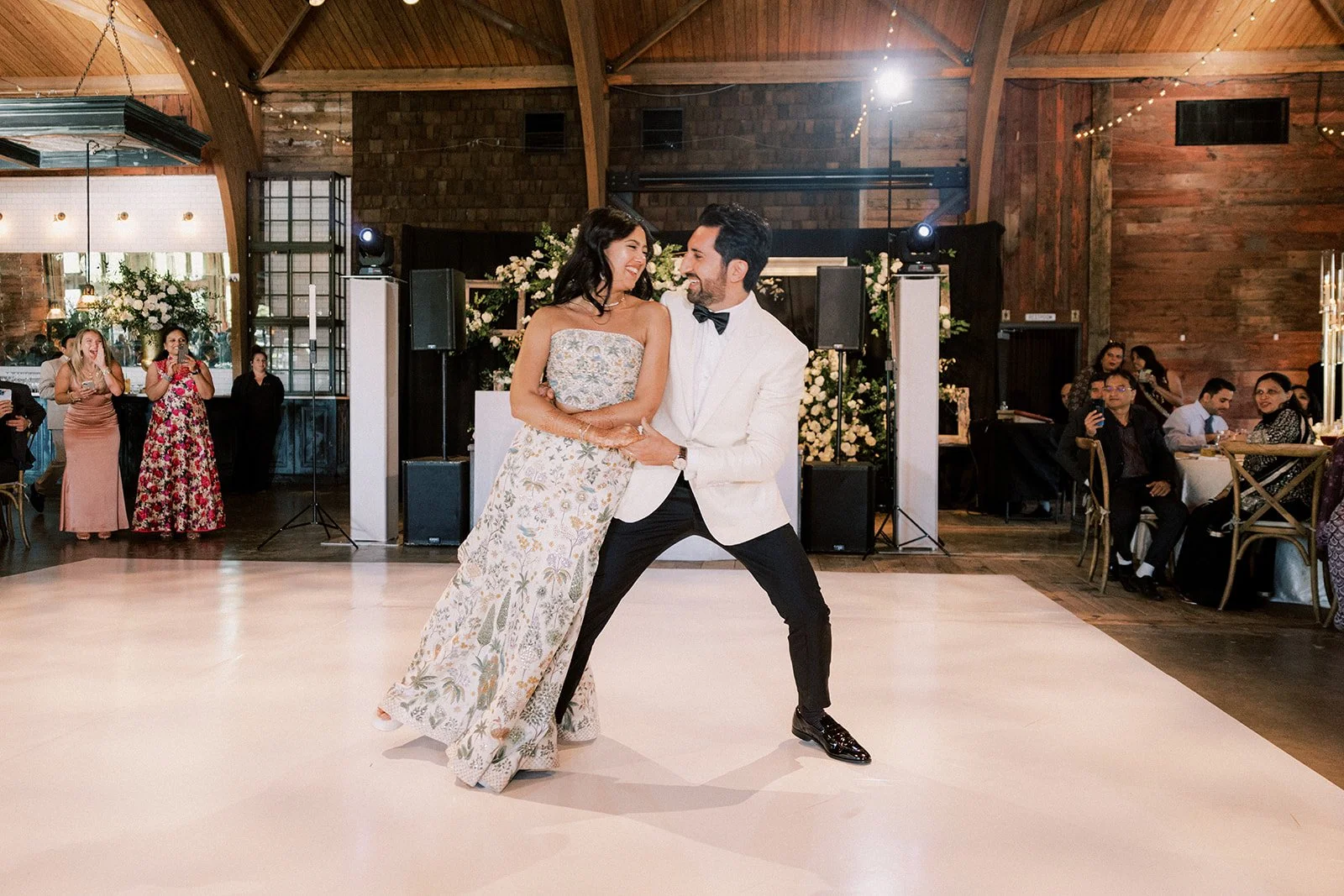 A couple dancing at a wedding reception, with guests seated at tables and taking photos in the background, in a rustic venue with wooden walls and string lights.