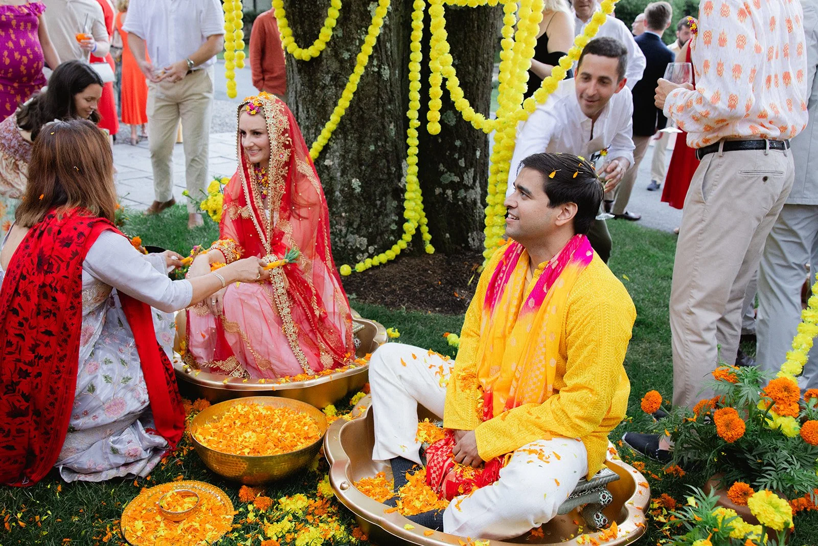 A traditional Indian wedding ceremony outdoors, with a bride in a red and gold sari and groom in yellow and white attire, sitting on a decorated platform surrounded by flower petals and garlands, while guests in colorful clothing celebrate around the