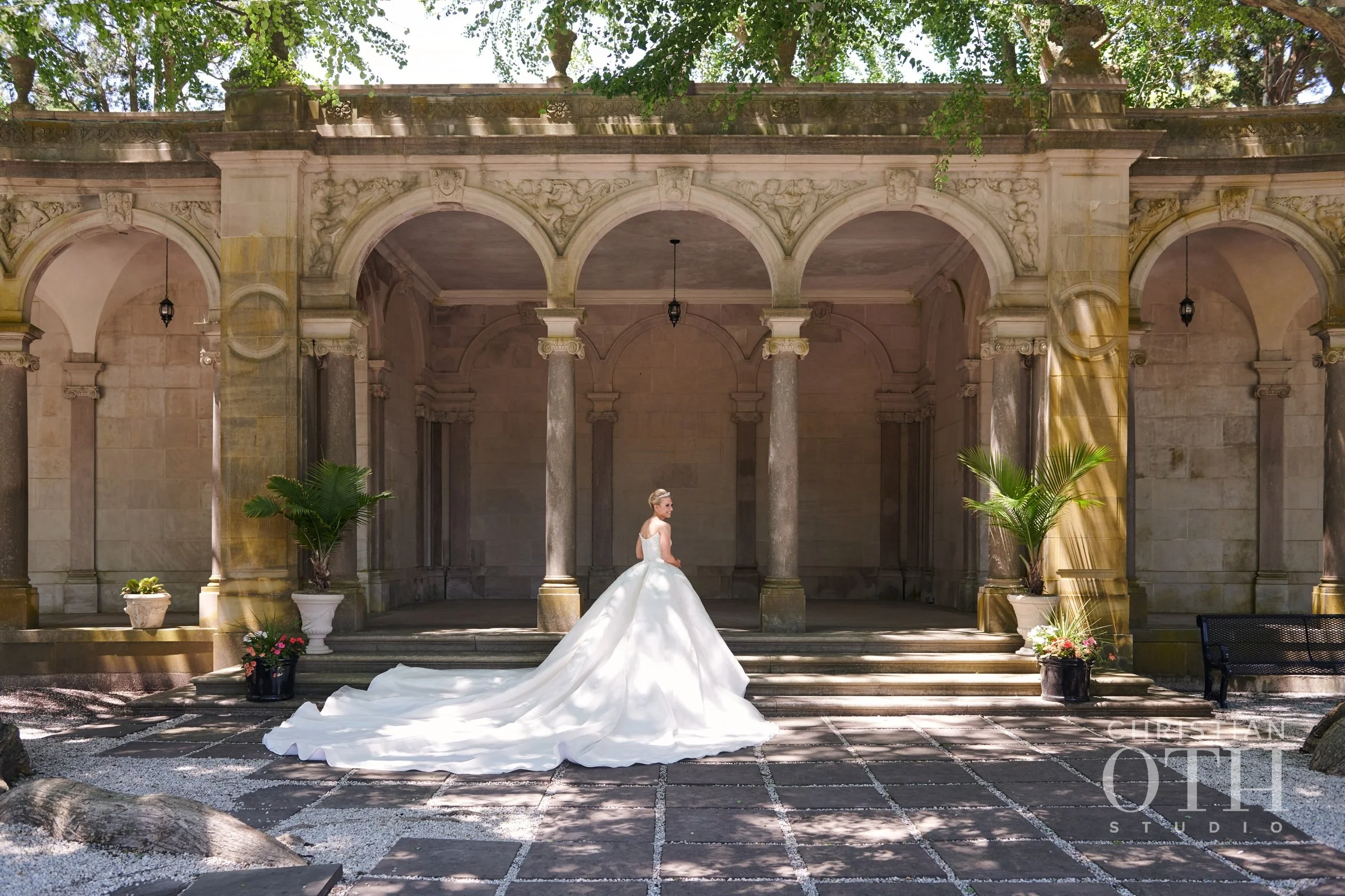 A bride in a white wedding dress with a long train standing on steps in front of an ornate stone archway with columns and greenery.