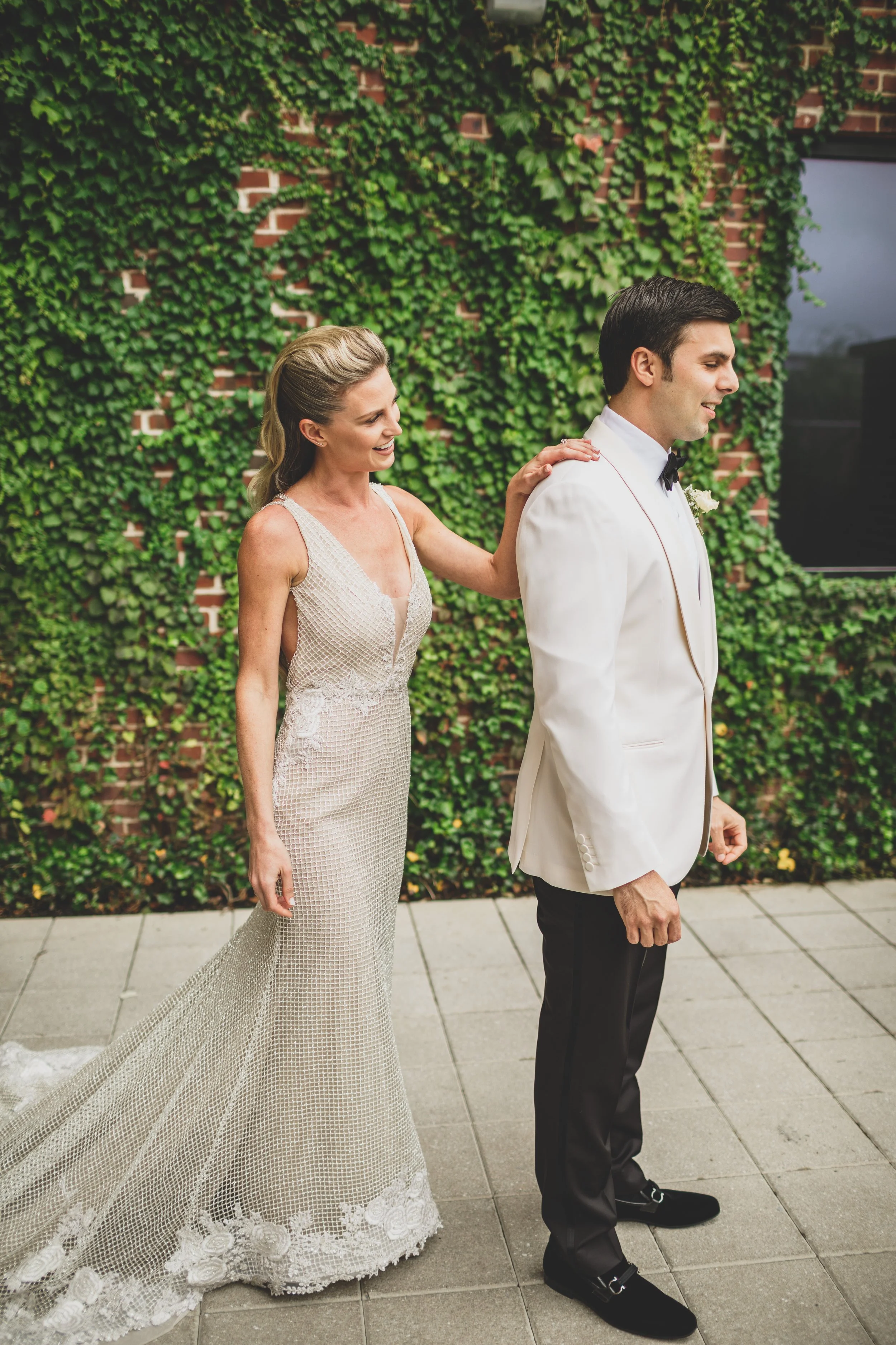 A bride and groom at a wedding, standing outdoors on a tiled patio with green ivy-covered brick wall in the background. The bride is smiling and gently touching the groom's shoulder.