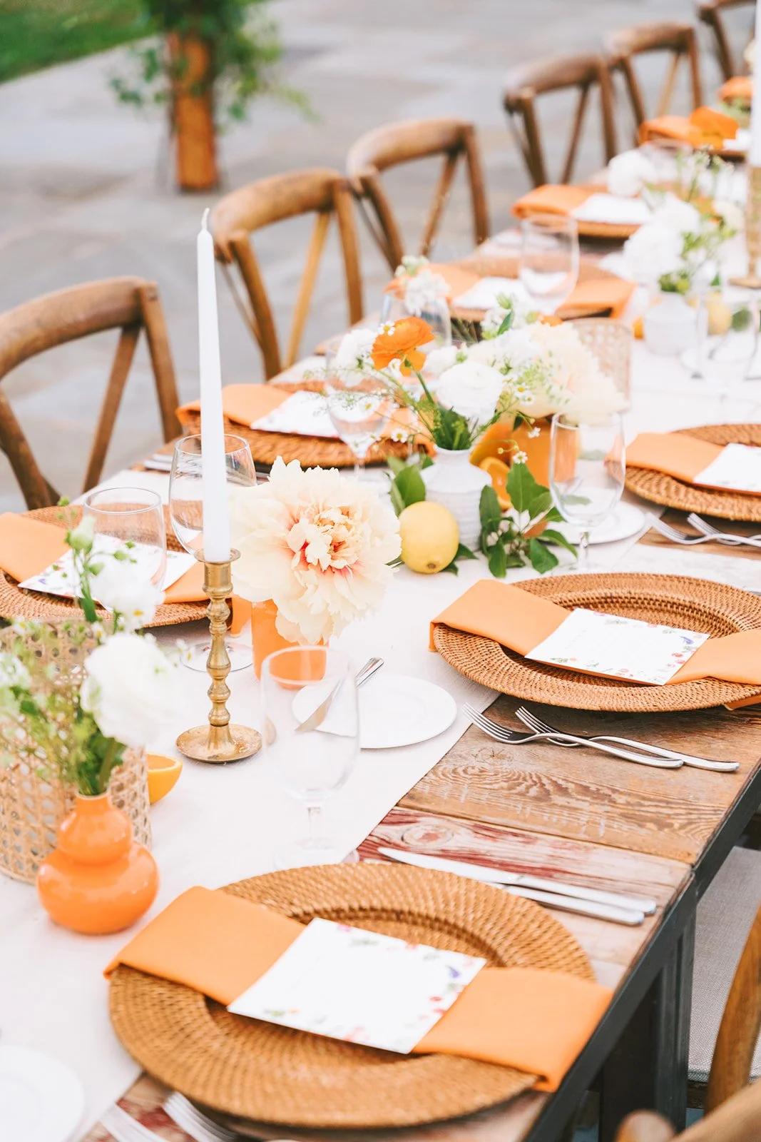 Beautiful outdoor dining table setup with orange and white floral centerpiece, candles, orange napkins, wicker placemats, and glassware surrounded by wooden chairs.