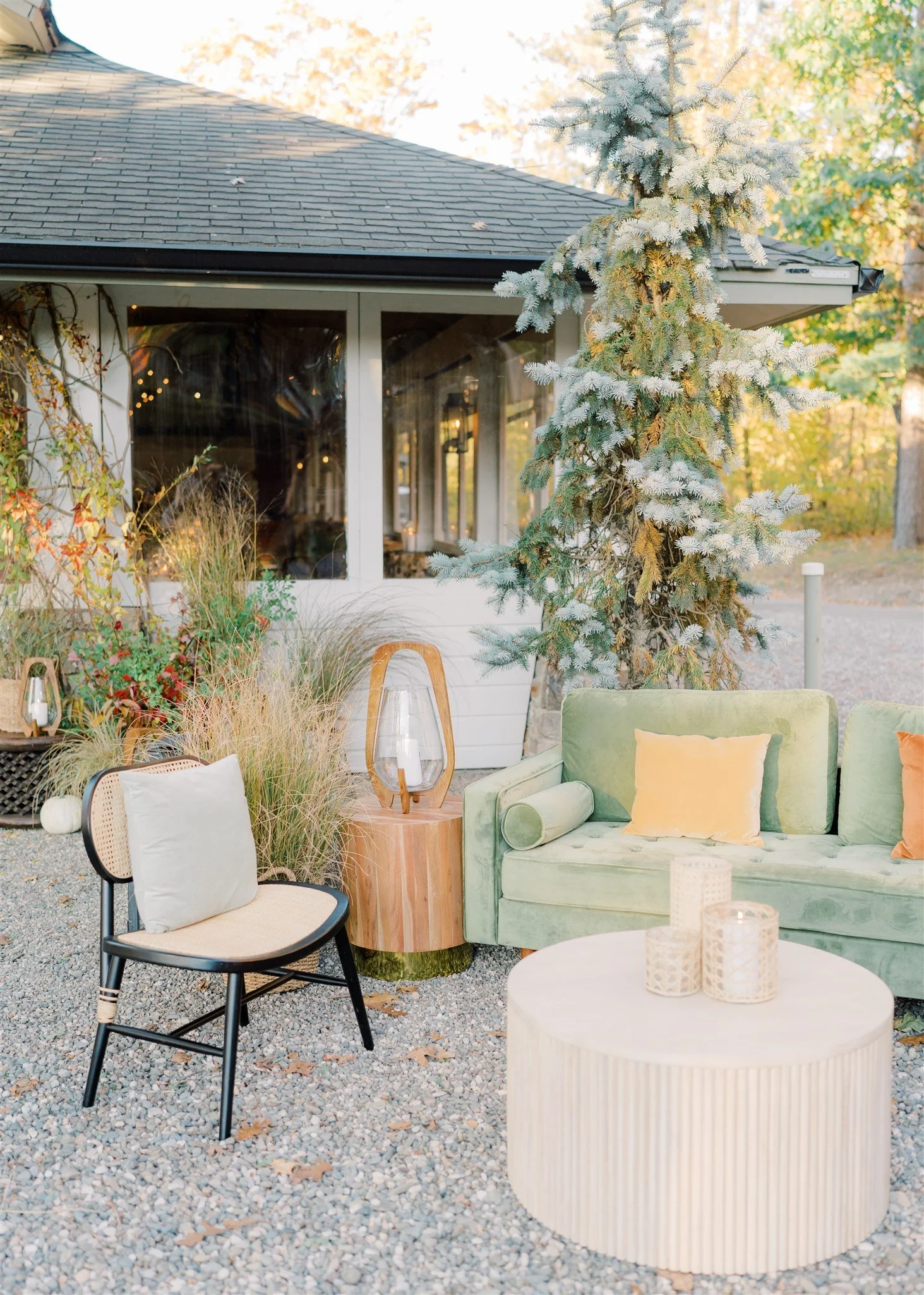 Outdoor patio with a green velvet sofa, a black chair with a cushion, a round white table with candles, and a large evergreen tree, with a house with screened porch in the background.