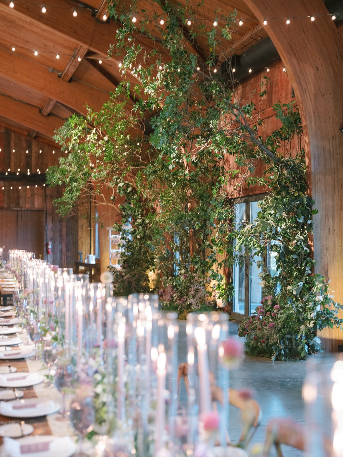 Elegant indoor wedding reception with a long table, floral centerpieces, and fairy lights, featuring a large indoor tree and wooden walls.