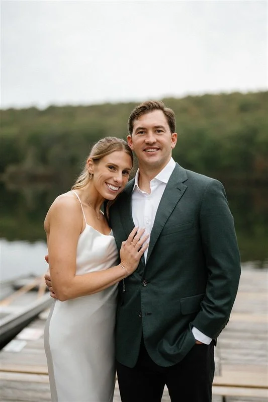 A smiling woman in a white dress and a man in a dark suit stand together outdoors near a lake with trees in the background.