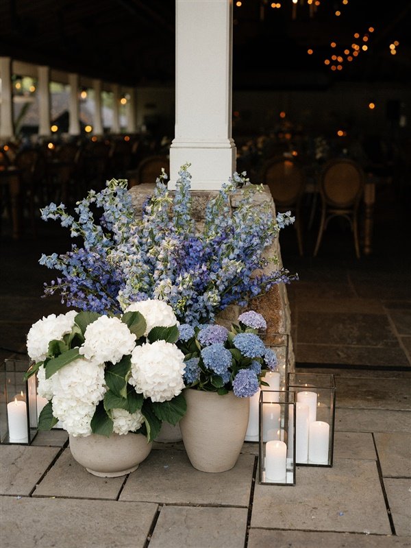 Decorative floral arrangement with white hydrangeas and blue delphiniums in beige pots, surrounded by white candles in glass holders near a white pillar in a dimly lit indoor setting.