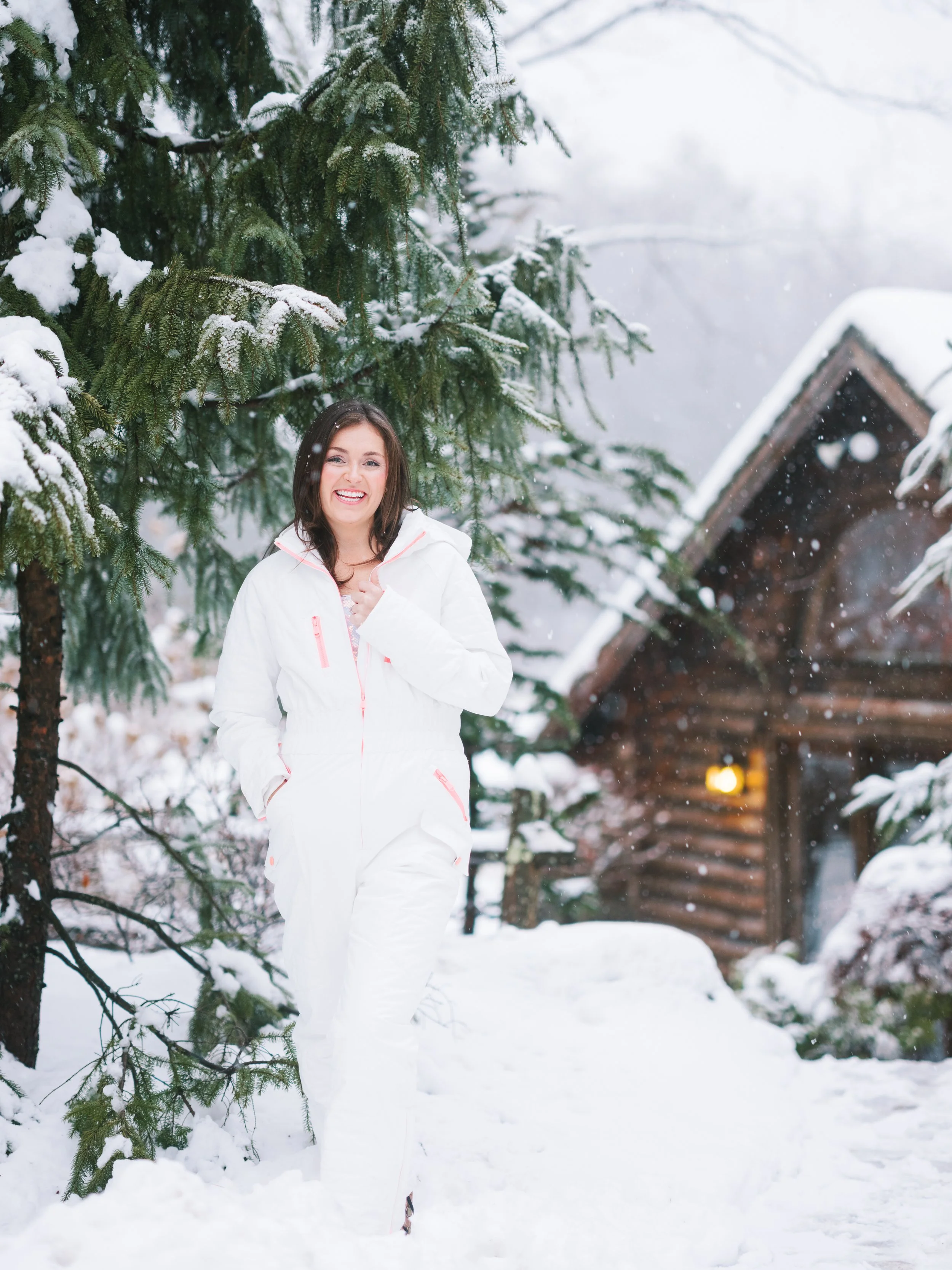 A woman dressed in a white winter jumpsuit walking through snow near a forest with snow-covered trees and a rustic cabin in the background, smiling.