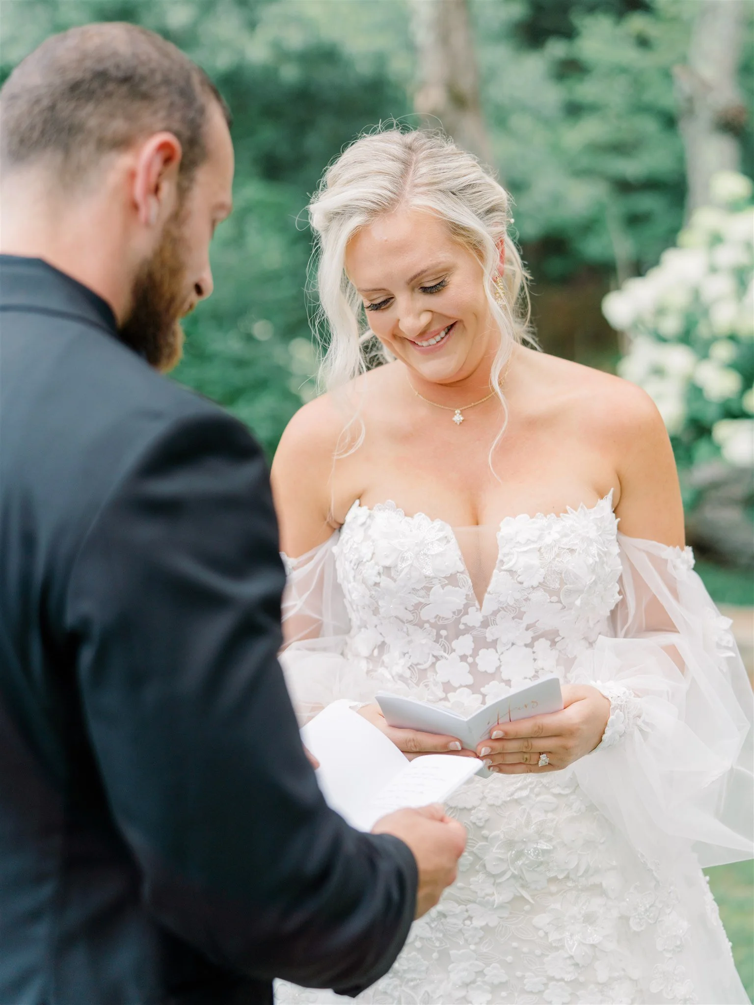 A bride and groom exchanging vows outdoors during a wedding ceremony, with the bride smiling and holding a small notecard.