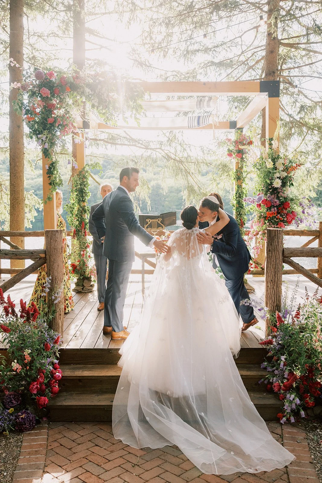 A wedding ceremony outdoors with a bride in a white gown and groom in a dark suit, surrounded by floral decorations and trees.