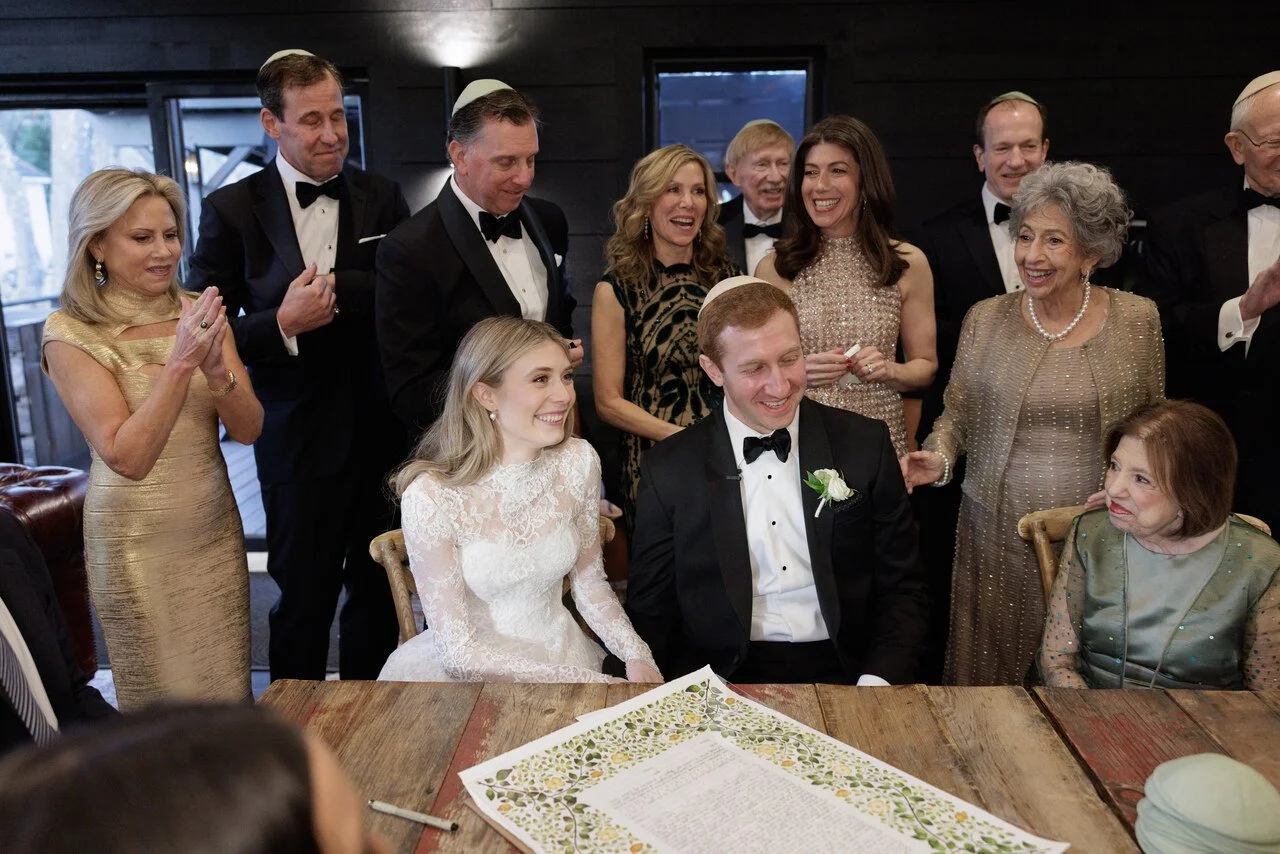 A wedding celebration with a bride and groom seated at a table, surrounded by family and friends dressed in formal attire, smiling and clapping.