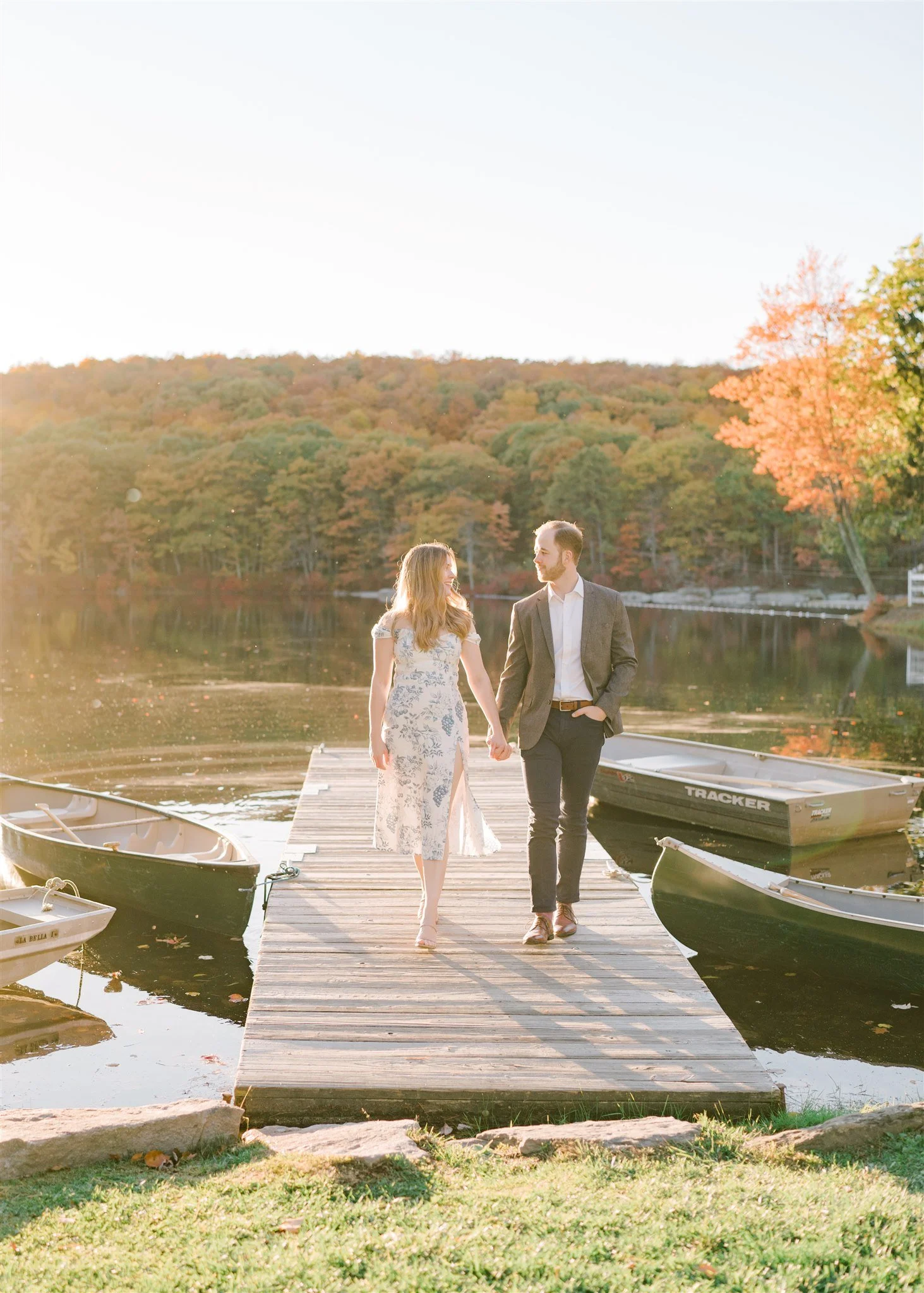 A couple walking hand in hand on a wooden dock by a lake, surrounded by fall foliage, with small boats docked nearby during golden hour sunlight.