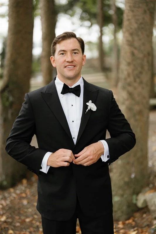 A man in a black tuxedo with a bow tie, white shirt, and a white flower boutonniere, standing outdoors with trees in the background.