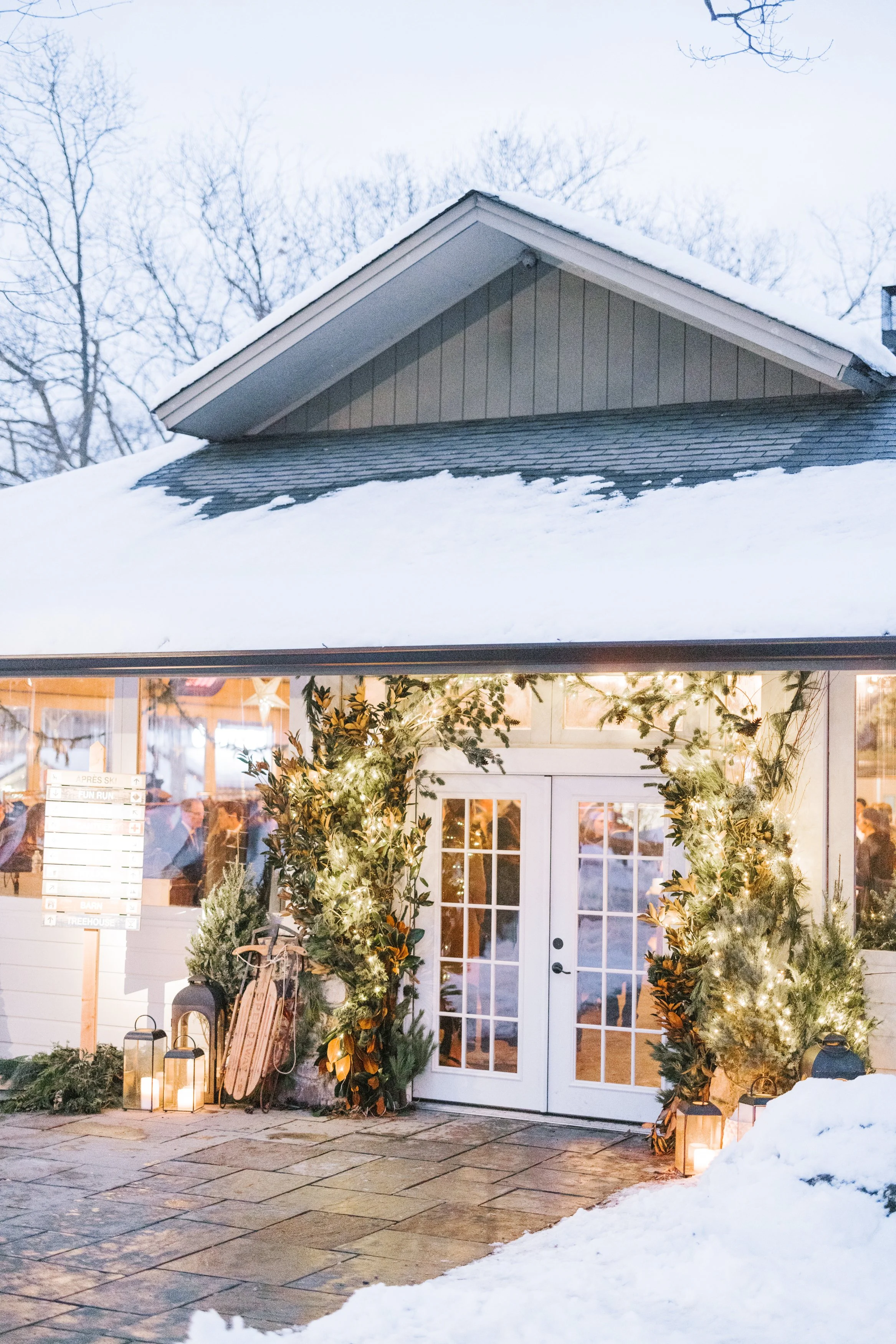 Decorated entrance of a building with twinkling lights, greenery, and lanterns, surrounded by snow, with visible people inside.